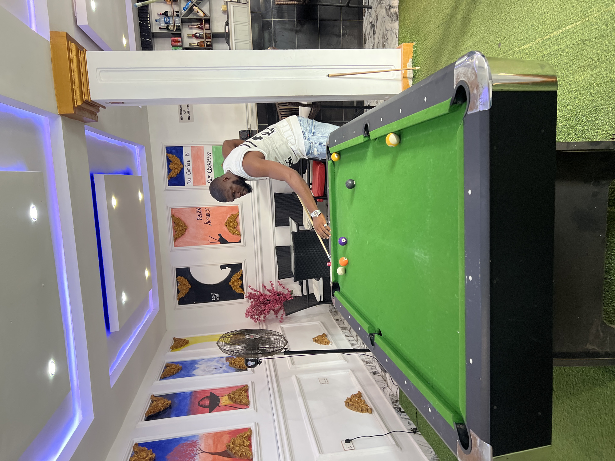 A man is playing billiards in a room. He is wearing a white tank top and ripped jeans. The room has a green billiard table with several balls on it, including a white cue ball, a yellow ball, and a purple ball. There are paintings on the wall, some with text that reads "Good night," "Relax Request," "Your Comfort is," and "Our Concern." There is also a fan in the room. The lighting in the room is provided by ceiling lights and some blue accent lighting. The room appears to be indoors, possibly a game room or a lounge area. The overall impression is a casual and recreational setting.