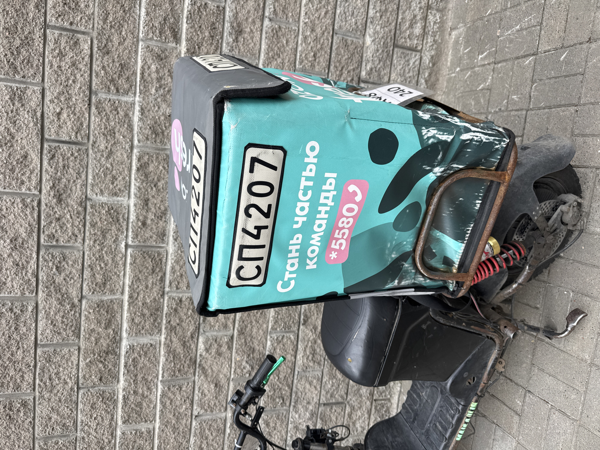 An electric scooter with a large, turquoise delivery box is parked on a paved surface against a grey brick wall. The delivery box has white text that reads "СП4207" and "Стань частью команды *5580". There is a pink circular logo with white text on the lid of the box. The scooter is black with red shock absorbers. The scene is located in Ladozhskoe Ozero, Russia.