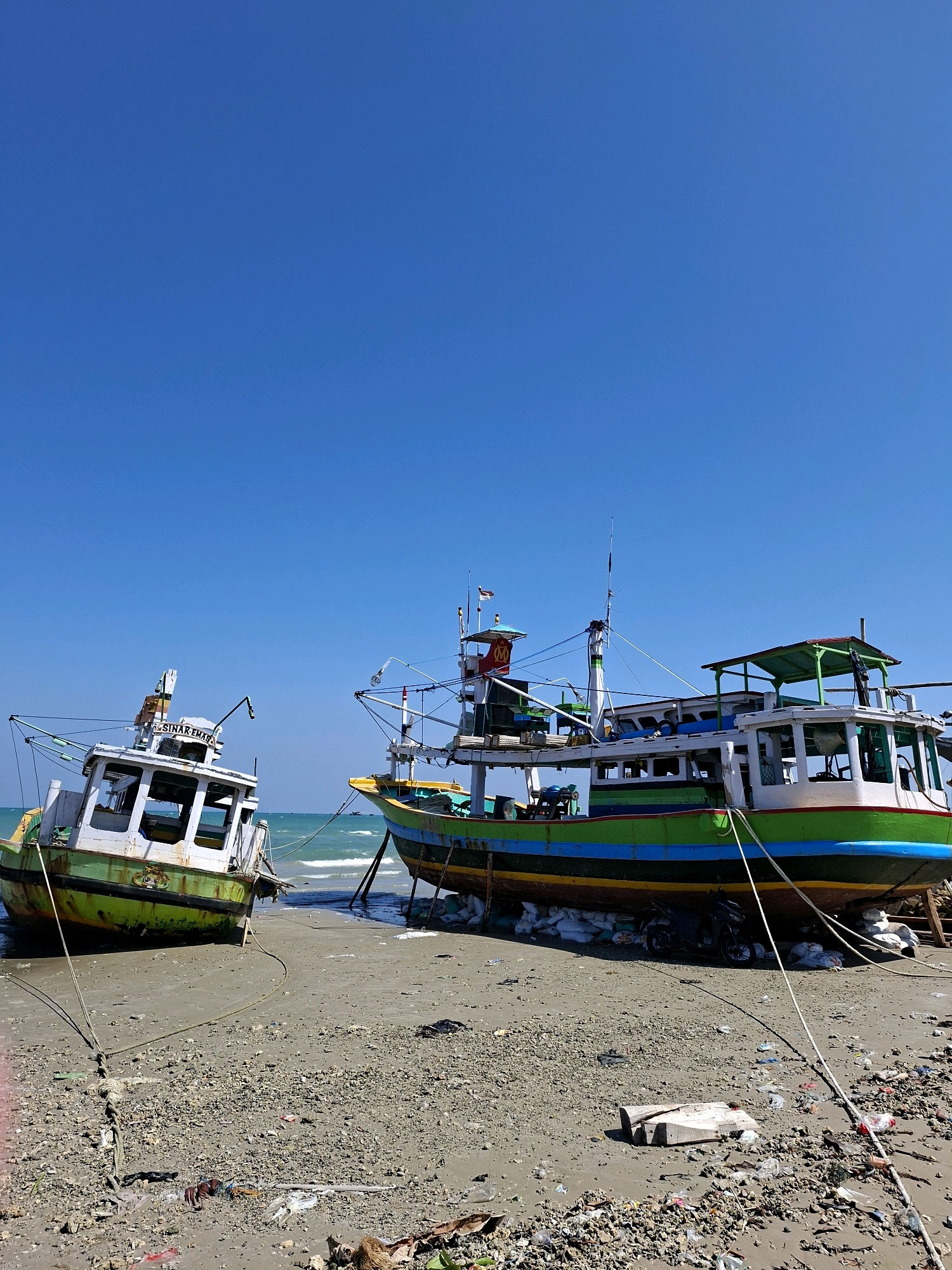 Two fishing boats are beached on the shore of Camplong, Indonesia, under a clear blue sky. The boat on the left, mostly white with a green hull, lists slightly to the left and has the name "SINAR EMAS" visible on its cabin. The boat on the right is larger and more colorful, with a blue, green, and yellow hull, and a multi-level cabin structure. Both boats appear to be out of the water, resting on the sandy, debris-strewn beach. Ropes are secured from the boats to the shore. The water of the ocean is visible behind the boats, with small waves and a few distant boats on the horizon. The beach is littered with various types of trash, including plastic bottles, bags, and wood debris.