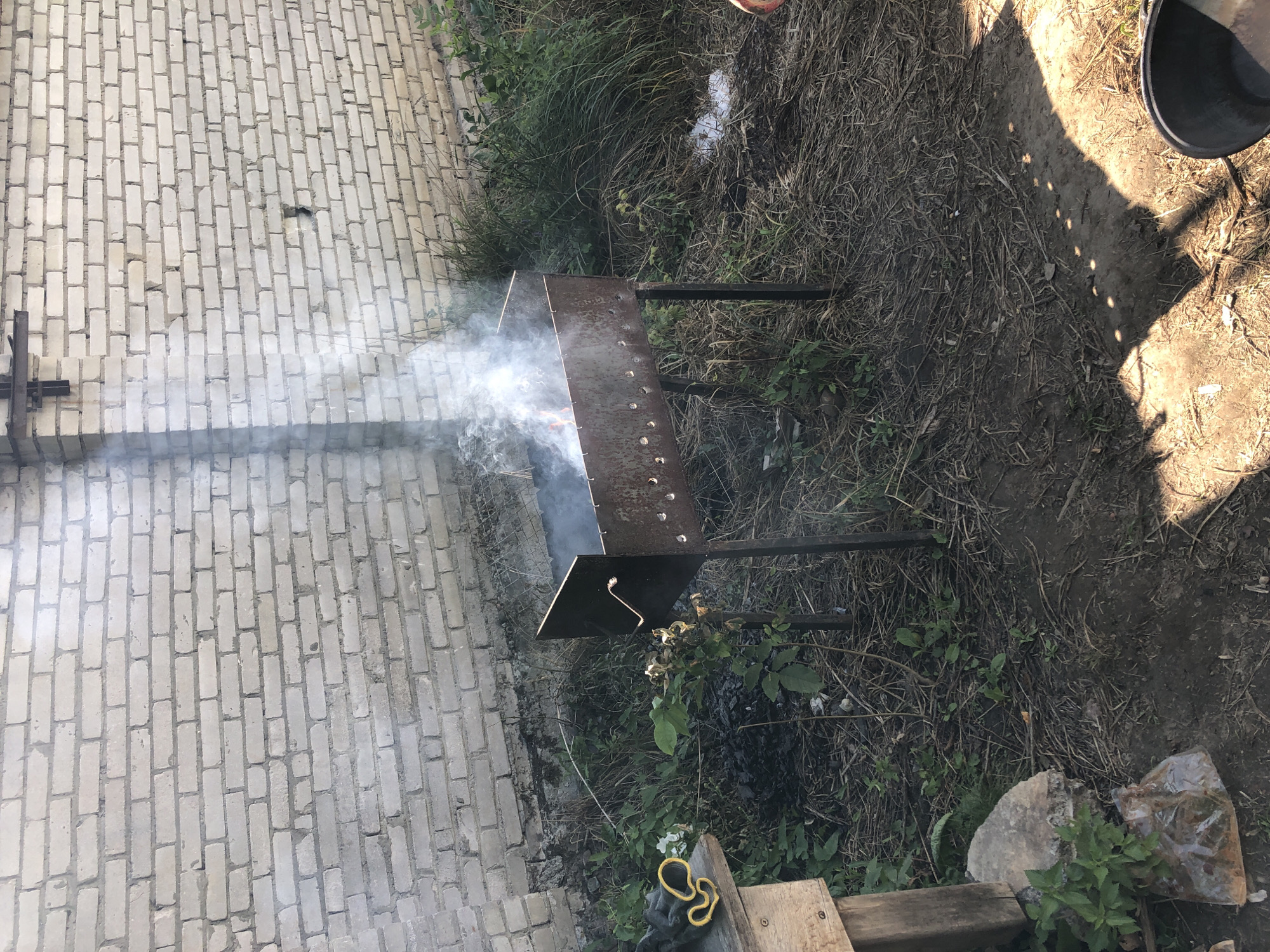 In Lviv, Ukraine, a metal barbecue grill, known locally as a "mangala," is situated outdoors. Smoke billows from the grill, indicating active use. The grill rests on a patch of dry grass and dirt, with a worn wooden table to its left. A white brick wall forms the backdrop of the scene. The ground is littered with dried grass, twigs, and debris. Sunlight casts dappled shadows on the ground, suggesting it is daytime. The overall impression is of a casual, outdoor cooking setup.