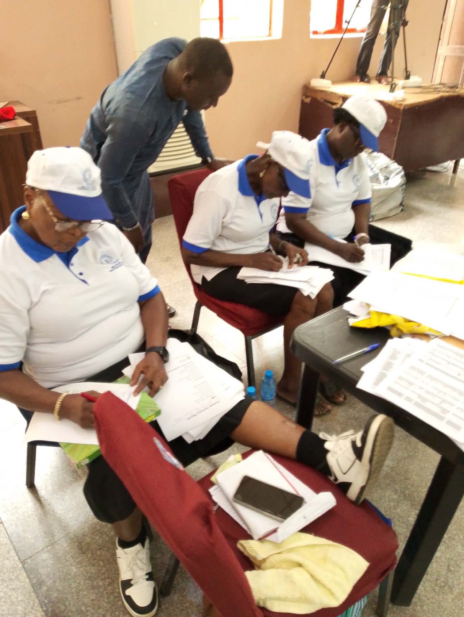 In an indoor setting in Kubwa, Nigeria, four individuals are engaged in an activity involving paperwork, likely a training session, registration, or administrative task. Three women are seated, wearing matching white polo shirts with blue collars and trim, and white caps with blue visors, each adorned with a circular logo containing text and a graphic. They are focused on papers spread on their laps and on a dark table to their right, some writing with pens.
The woman in the foreground wears eyeglasses, a gold-colored bracelet, and a wristwatch, with her leg extended showing a white and black sneaker. Beside her, on a red fabric surface, lies a black smartphone and a folded yellow cloth. Another woman to her right is also writing, with a blue plastic water bottle visible on the floor near her bare feet. The third woman in uniform is partially obscured behind the second, also appearing to be engaged with documents.
A man, dressed in a blue long-sleeved garment, stands leaning over from the left, observing or interacting with the seated women. The room has light-colored walls and windows, indicating daytime. In the background, a stage or raised platform is visible with a camera tripod, suggesting the event might be recorded or documented. Papers are scattered on the table and floor, reflecting an active work environment.