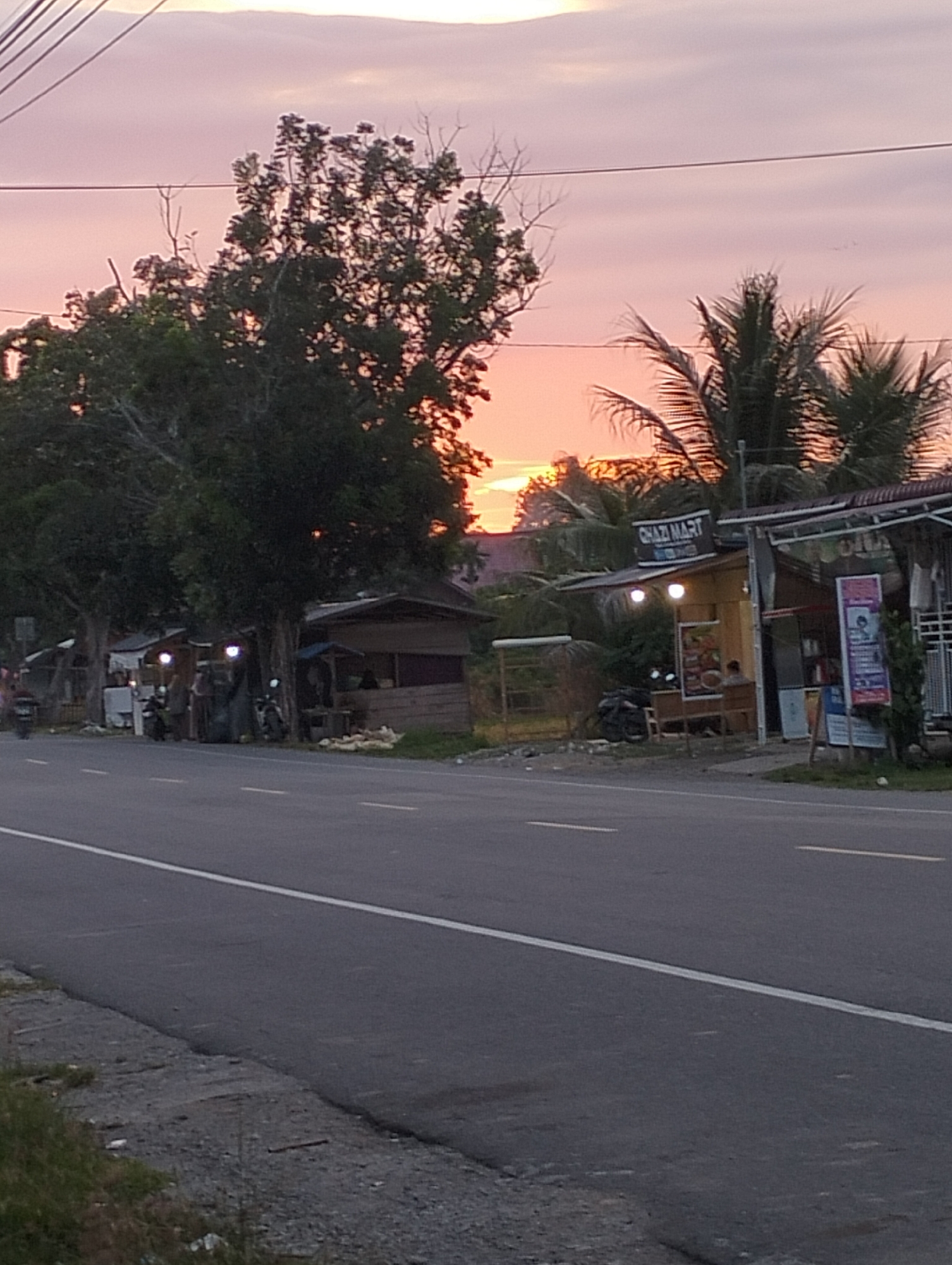 In Blangmetimu, Indonesia, at dusk, a roadside scene unfolds with a paved road in the foreground, marked with white lines. To the right of the road, a row of small wooden stalls or shops are visible. One stall has a sign that reads "GHAZI MART". There are motorcycles parked near the stalls, and a few figures are present, appearing to be customers or vendors. Tall trees with dense foliage dominate the left side of the image, partially obscuring the stalls and the sky. The sky exhibits the soft, pastel hues of a sunset, with streaks of pink and orange. Palm trees are visible behind the stalls, adding to the tropical atmosphere. The overall impression is a quiet evening scene in a developing area.