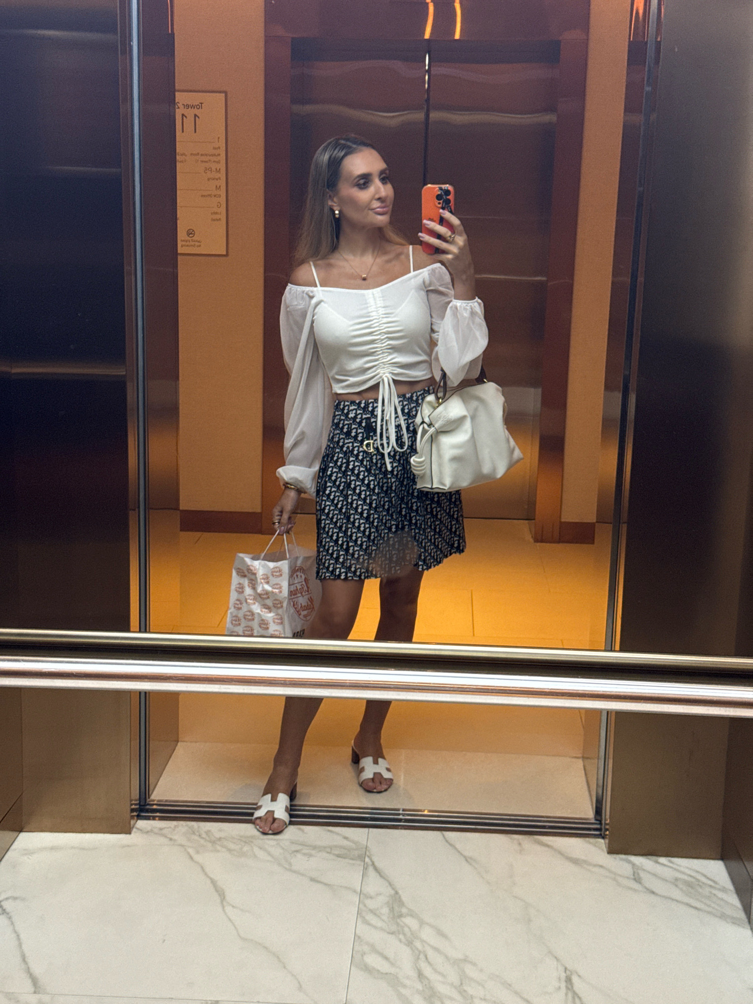 In an elevator in Dubai, United Arab Emirates, a woman stands taking a mirror selfie. She is wearing a white off-the-shoulder crop top with ruched detailing and long sleeves, a dark navy skirt with a white logo pattern, and white slip-on sandals. She is holding a white handbag in her left hand and a white shopping bag in her right hand. The elevator has mirrored walls and a marble floor. A sign is visible on the wall behind her.