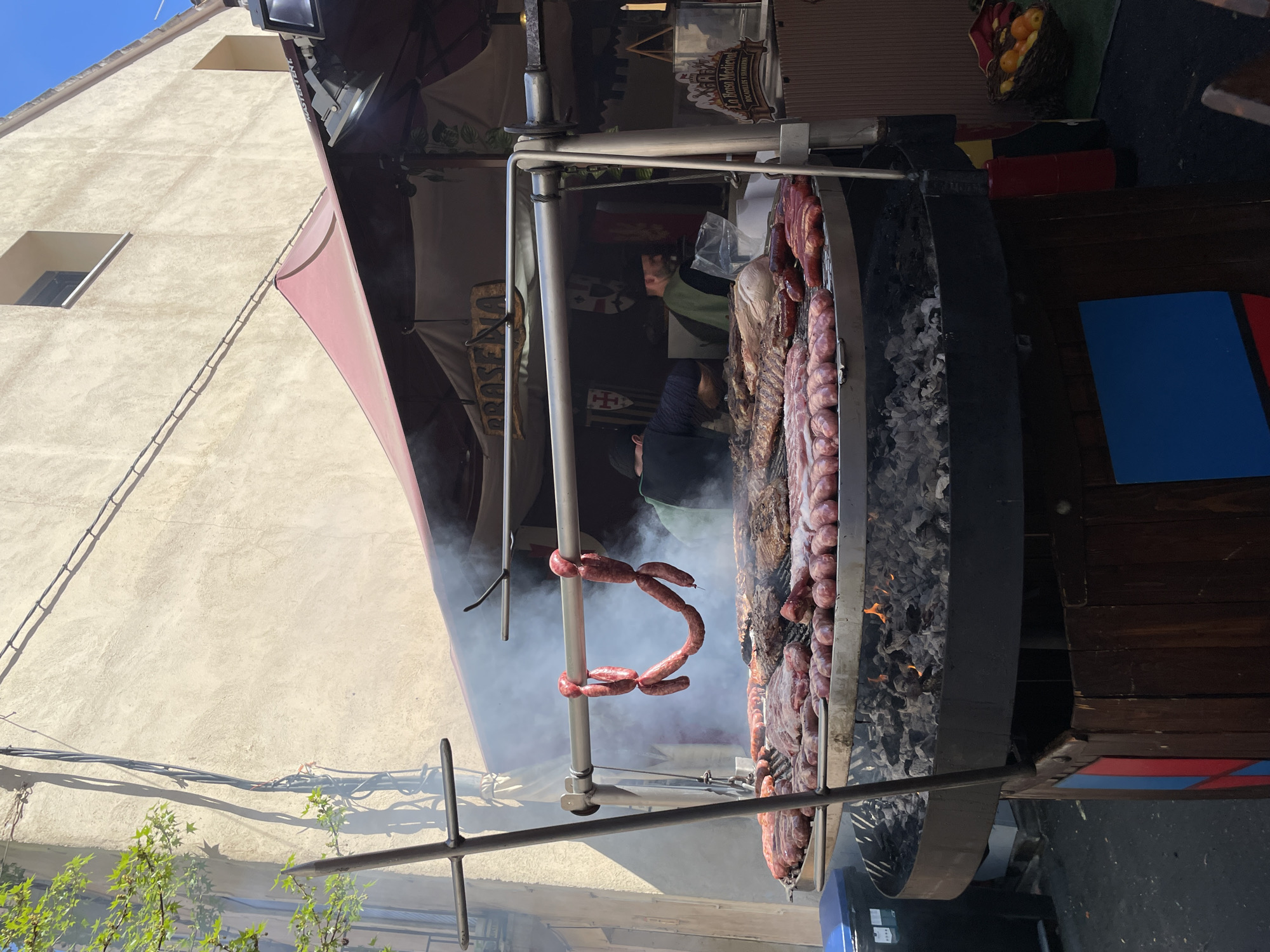 The image depicts a medieval-style food stall in Hostalric, Spain. Sausages are hanging on a metal pole. A large grill behind the pole is filled with various meats cooking over hot coals. Smoke rises from the grill and food. People are visible behind the stall's counter. Buildings are in the background.