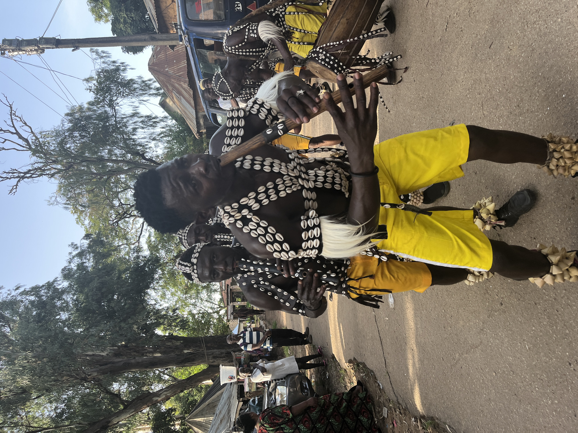 This image captures a vibrant cultural event in Jos, Nigeria. The central subjects are a group of men adorned in traditional attire, likely participating in a performance or ceremony. They are dressed in yellow shorts and elaborate necklaces made of white shells. Some individuals are playing musical instruments, including a flute-like instrument and a drum. Their faces show focused expressions, suggesting engagement in the activity.
The setting appears to be an outdoor public space, possibly a street or a market area, indicated by the asphalt ground, a vehicle, and a building with a corrugated iron roof in the background. Trees with lush green foliage frame the scene, and the bright sunlight suggests it is daytime. The overall atmosphere seems lively and festive.
Notable details include the intricate shell adornments on the performers, the traditional musical instruments, and the vibrant yellow of their shorts. The presence of other people in the background, dressed in more casual clothing, hints at a community gathering. There is no visible text in the image. The scene conveys a sense of cultural pride and celebratory spirit.