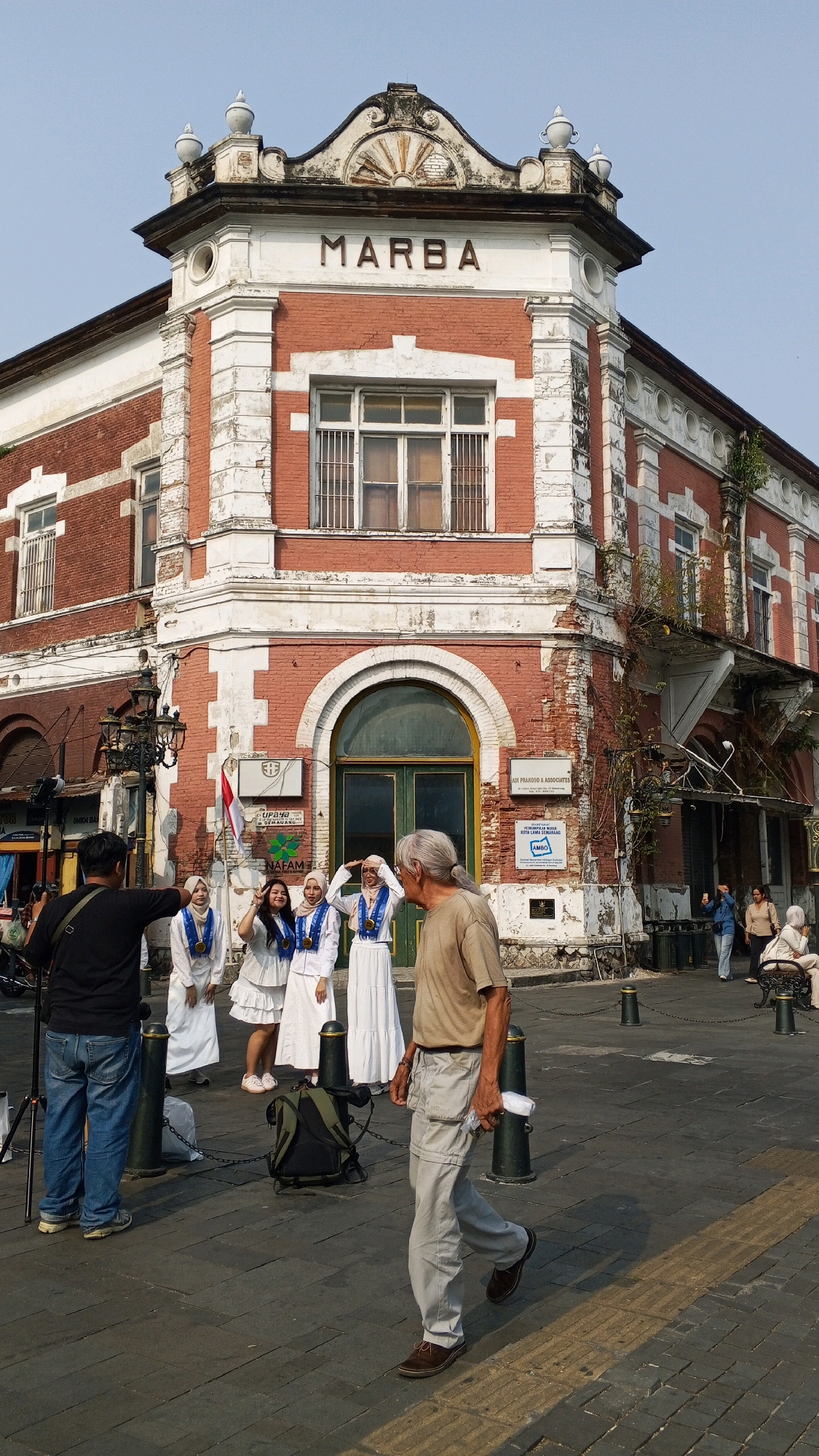 The image shows the iconic Marba Building in Semarang, Indonesia, a historic structure with red brick and white architectural details. The building is situated in a public square, likely Kota Lama Semarang, known for its colonial-era architecture.
In the foreground, a man wearing a dark shirt and jeans stands behind a tripod, appearing to be a photographer. He is facing a group of four young women dressed in white gowns and headscarves, adorned with blue sashes and medals. They are posing for the camera, with two of them making hand gestures.
To the right of the group, an older man with gray hair and a beige t-shirt walks away from the camera. He is wearing light-colored pants and dark shoes, and is holding a white cloth in his right hand.
The square is paved with brickwork, and several dark bollards with chains are visible, marking pedestrian areas. In the background, other people are seen in the distance, some sitting on benches and others walking. The sky is clear and blue. The overall scene depicts a moment of cultural tourism and photography in a historically significant urban setting.