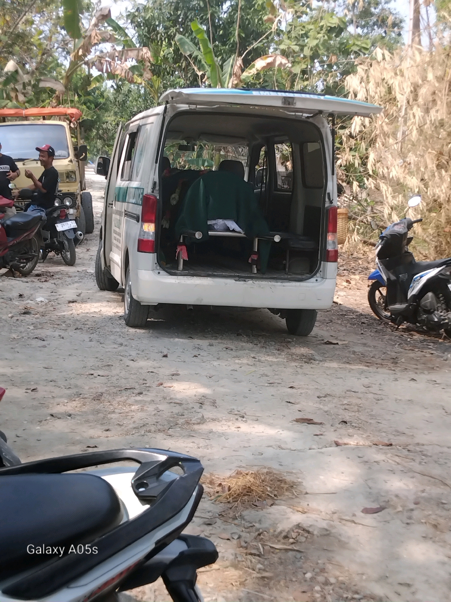 In Karanganyung, Indonesia, an ambulance van is parked on a dirt road lined with dry vegetation and trees. The rear doors of the van are open, revealing a stretcher with a green tarp covering its contents. To the left of the ambulance, a beige truck is visible, with two men standing nearby, one wearing a red cap. Two motorcycles are parked in the foreground, one red and one blue and white. The scene appears to be outdoors, possibly in a rural or semi-rural setting.