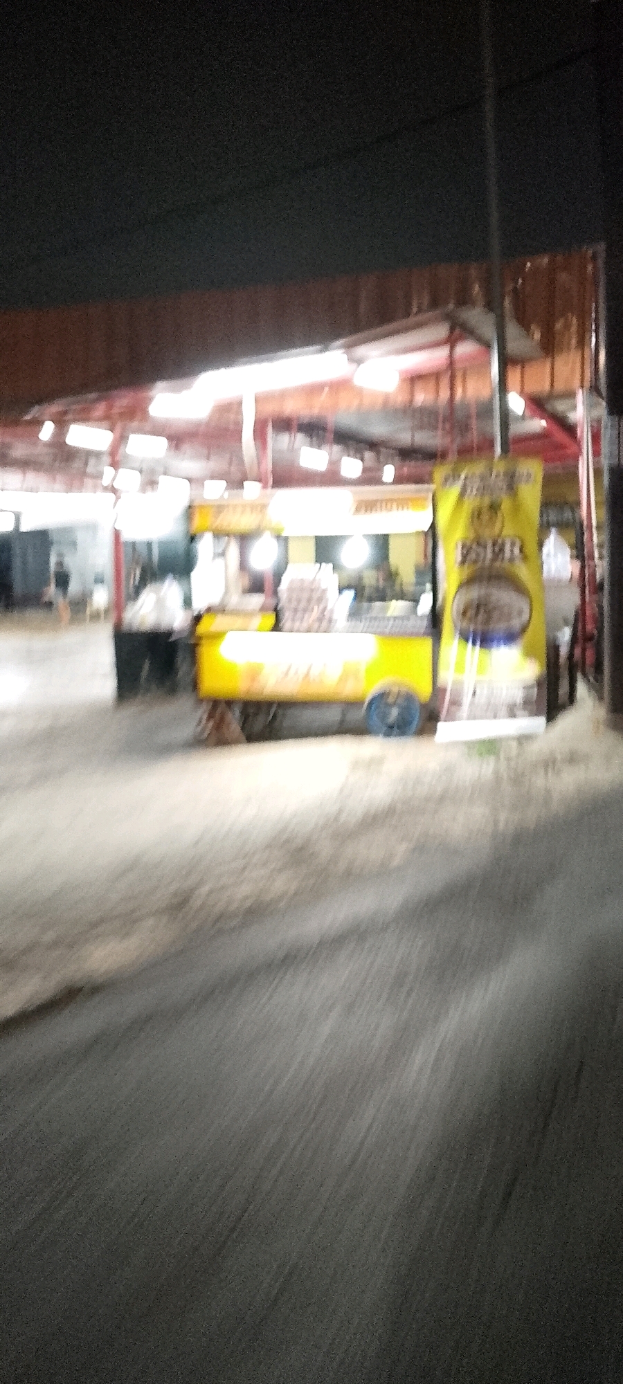 In East Jakarta, Indonesia, a nighttime scene depicts a street lined with food stalls. The focal point is a bright yellow food cart, possibly selling beverages or snacks, adorned with a banner featuring text in Indonesian, including "ESPER" and an emblem. The cart has blue wheels and is illuminated by overhead lights. Behind it, other stalls are visible, with a person walking in the background. The environment appears to be an open-air market or a commercial street. The foreground shows a blurred asphalt road, suggesting motion or low light conditions. The overall impression is that of a vibrant street food scene at night.