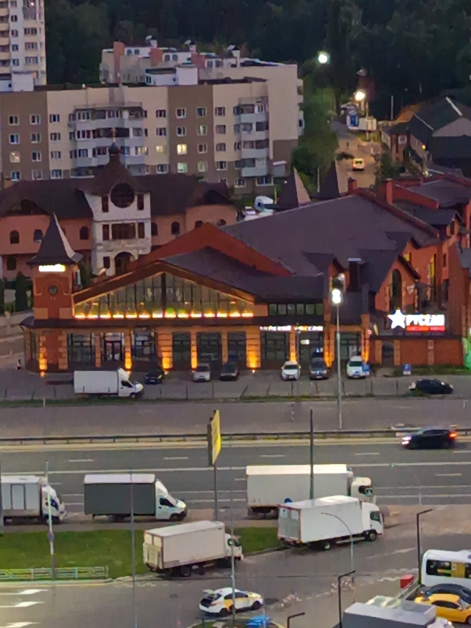 The image captures an aerial view of a street scene in Ostrovtsy, Russia, at dusk. In the foreground, a multi-lane road is visible with several white delivery trucks and a few cars. One white truck is parked near a sidewalk with a yellow sign. Further down the road, another white truck is moving, and a dark-colored car is also in motion.
Behind the road, a building with a red-tiled roof and numerous windows is prominently featured. This building appears to be a commercial establishment, with illuminated signage that includes a white star and Russian text. The building has a distinctive tower-like structure on its left side with a rounded roof and a circular window.
In the background, a large, multi-story residential building with a cream-colored facade and numerous windows is visible. Further in the distance, more buildings and trees can be seen, suggesting a mixed urban and suburban environment. The overall lighting suggests it is either early evening or late afternoon.