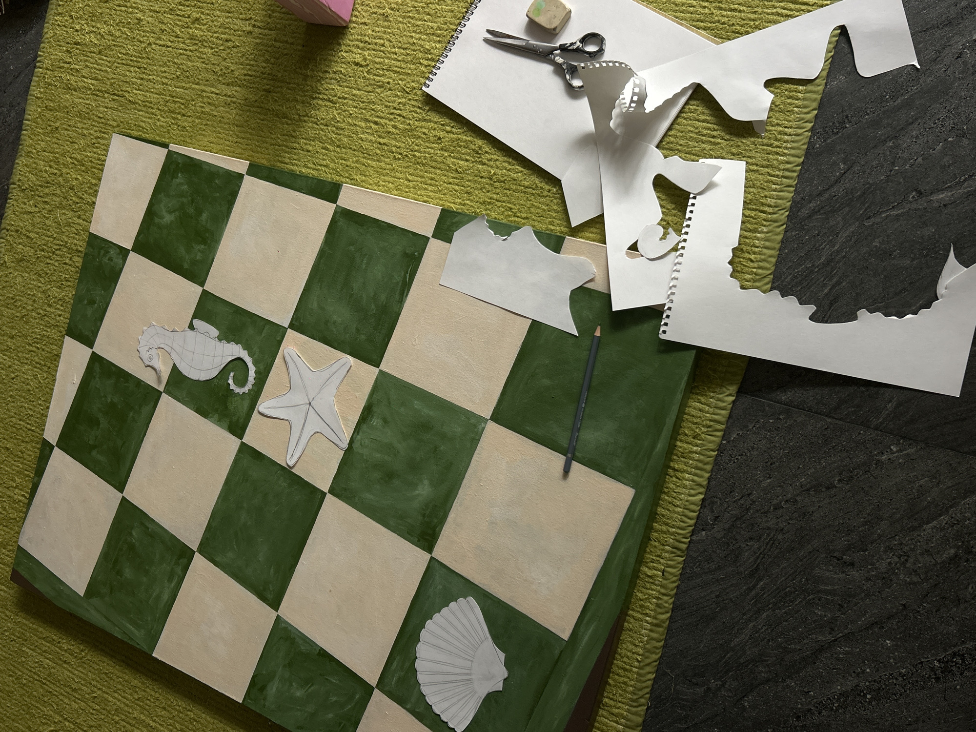 The image shows a scene on a green shag rug. A checkerboard with alternating green and beige squares sits on the rug. Paper cutouts of sea creatures including a seahorse, starfish, and seashell are placed on the board. To the right of the checkerboard, on the rug, is a notepad with loose leaf paper, scissors, and an eraser. A pencil rests on the checkerboard.
The location is not ascertainable from the image. Therefore, a description of North Kuta, Indonesia, is not possible.