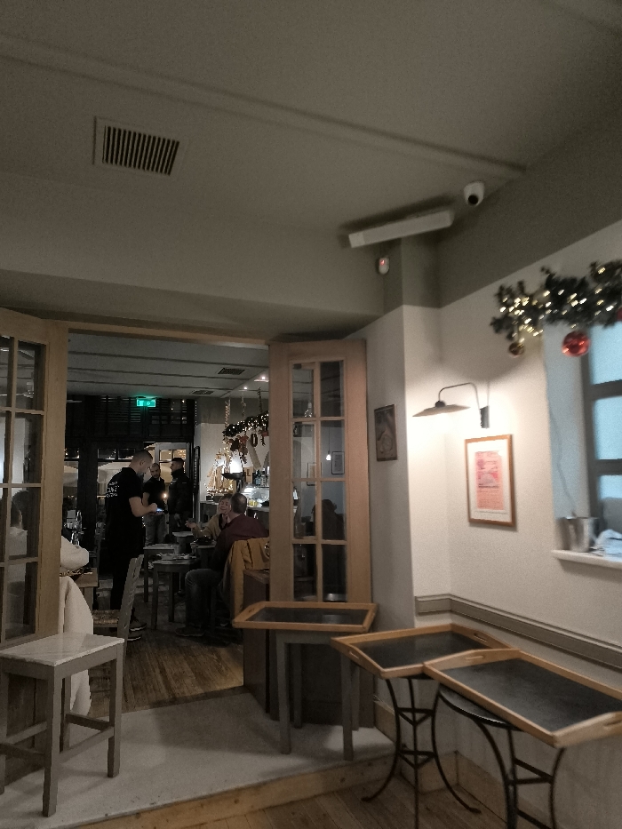 This image captures the interior of a cozy restaurant or cafe, likely during the evening, suggested by the warm, dim artificial lighting. The perspective is from a waiting area looking into the main dining space through open, light-colored wooden double doors with glass panes.
In the foreground, several small, square tables with dark tops and light wooden or metal frames are arranged, along with a simple wooden stool. The ceiling above features an air vent, a white rectangular light fixture, and a small white security camera. On the right-hand wall, a wall-mounted light fixture illuminates two framed pictures, one appearing to be a poster. A Christmas garland, decorated with lights and red ornaments, hangs above a window, on the ledge of which sits a silver bucket.
Through the open doors, several people are visible. A staff member, wearing a black shirt, is standing and interacting with a customer. Further into the room, a couple is seated at a table, appearing relaxed and engaged in conversation. The background shows glimpses of a bar area with bottles and glasses, and more tables with patrons. A green emergency exit sign is faintly visible in the distance.
The overall scene suggests a casual dining or social setting, with a warm and inviting atmosphere. The presence of Christmas decorations indicates the time of year is likely winter. No prominent text is clearly legible in the image, although a staff uniform and the emergency sign have text that is too small to decipher.