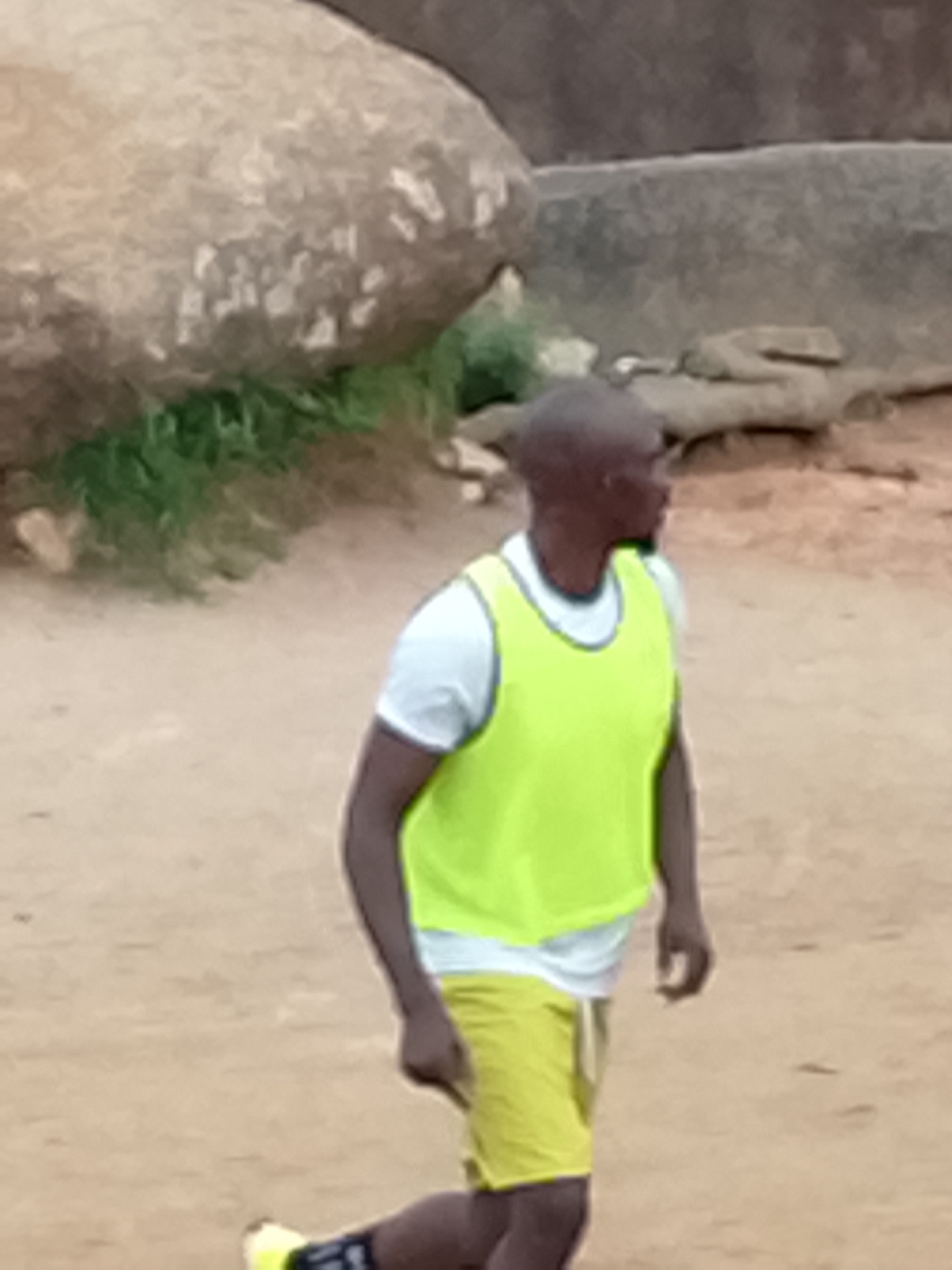 A person is walking on a dirt path in an outdoor environment. The person is wearing a white t-shirt, a bright yellow vest, and yellow shorts. The person's left foot is visible wearing a yellow shoe with black accents. In the background, there are large rocks and some sparse green vegetation. The scene appears to be in an undeveloped area, possibly on the outskirts of Jos, Nigeria, given the terrain and natural elements. The lighting suggests it is daytime.