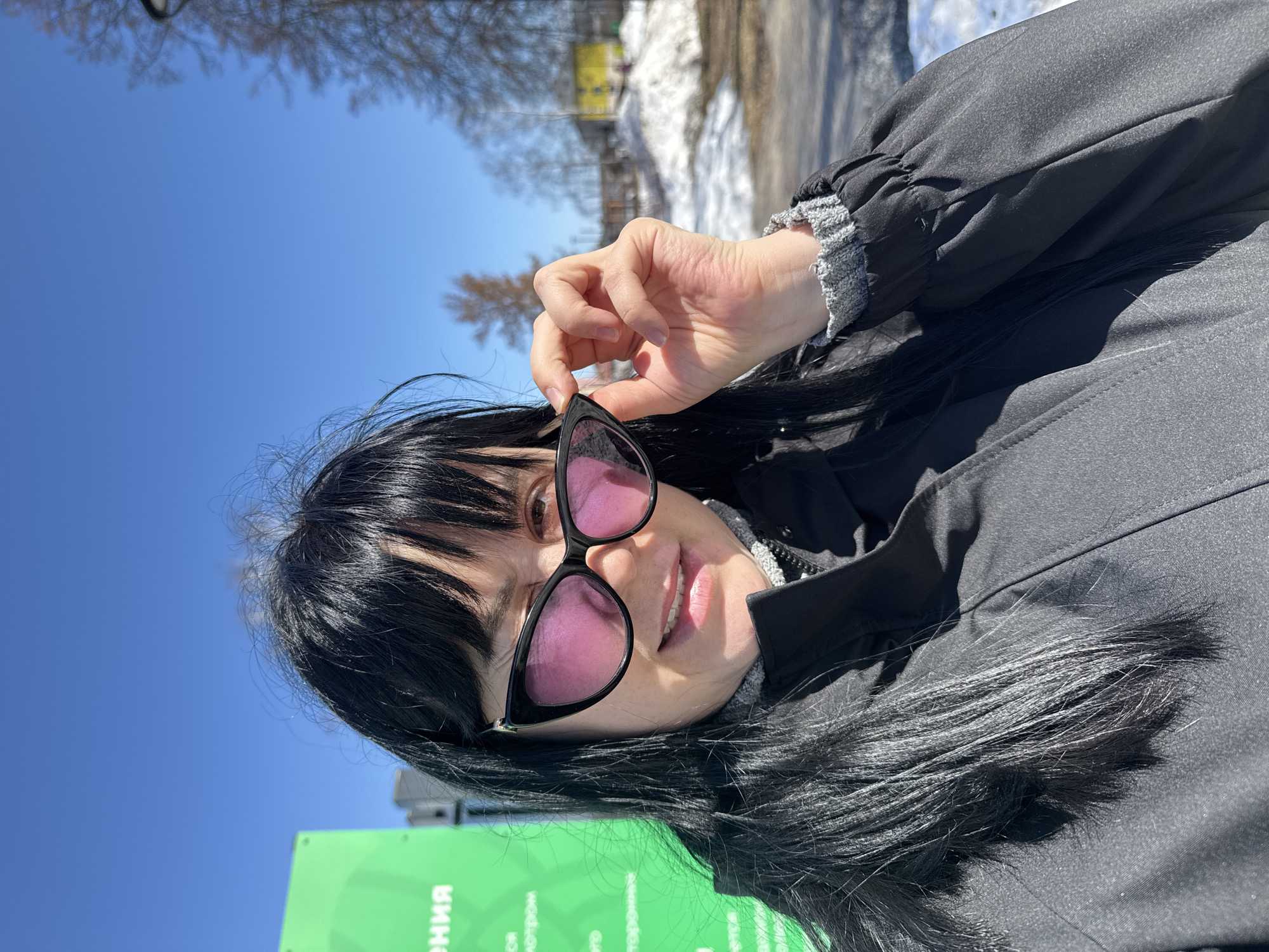 A woman with black hair and bangs holds up pink-tinted sunglasses. She is wearing a black jacket with a gray, textured sleeve cuff and a light-colored garment underneath. In the background, a blue sky is visible. A green sign with white writing and a patch of snow are partially visible. The photo was taken in Zav'yalovo, Russia.