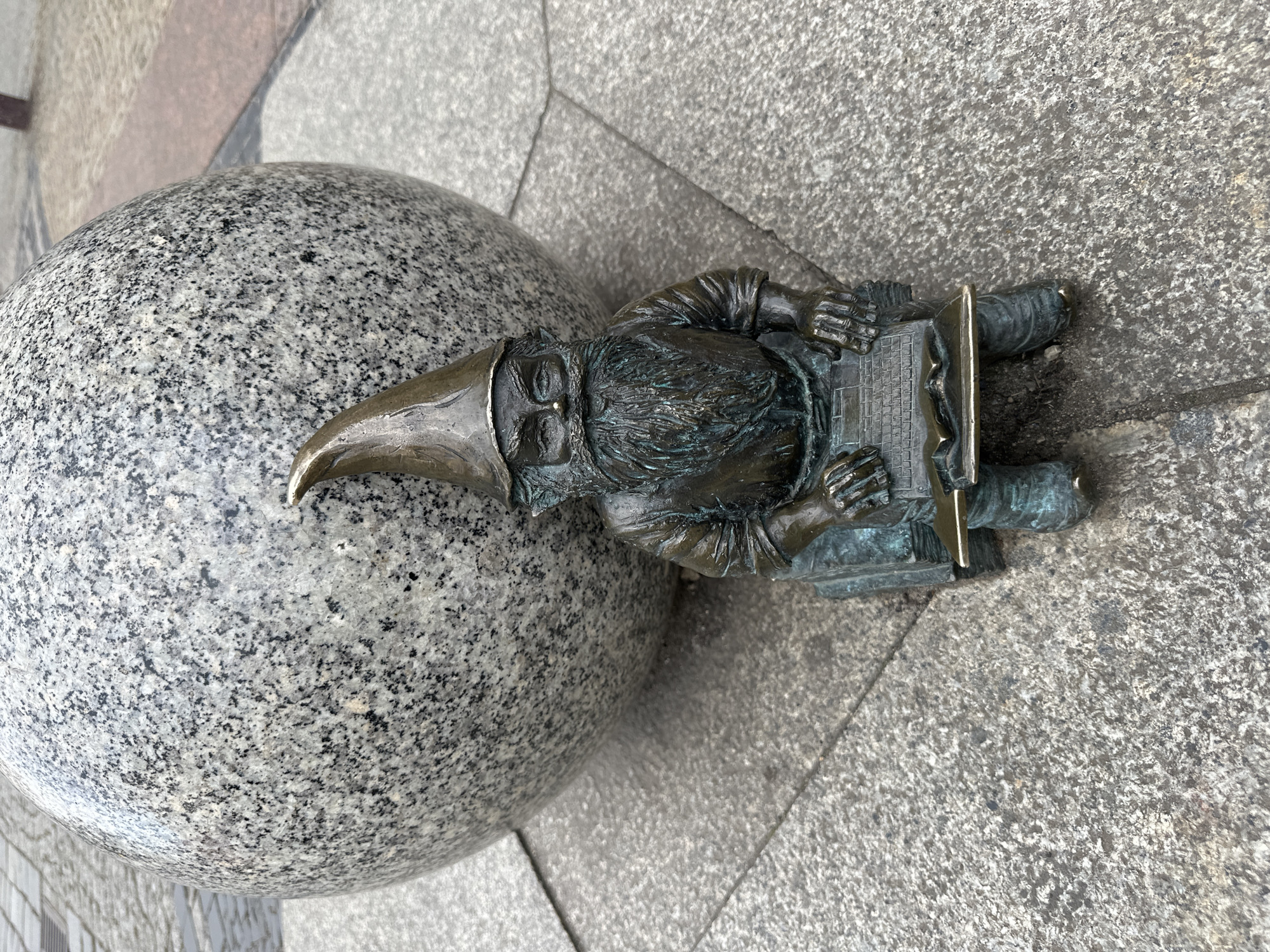 A bronze gnome statue is on a gray stone surface. The gnome wears a tall pointed hat and is holding a laptop. Beside the gnome is a gray stone sphere. This image depicts one of the famous Wrocław gnomes in Wrocław, Poland.