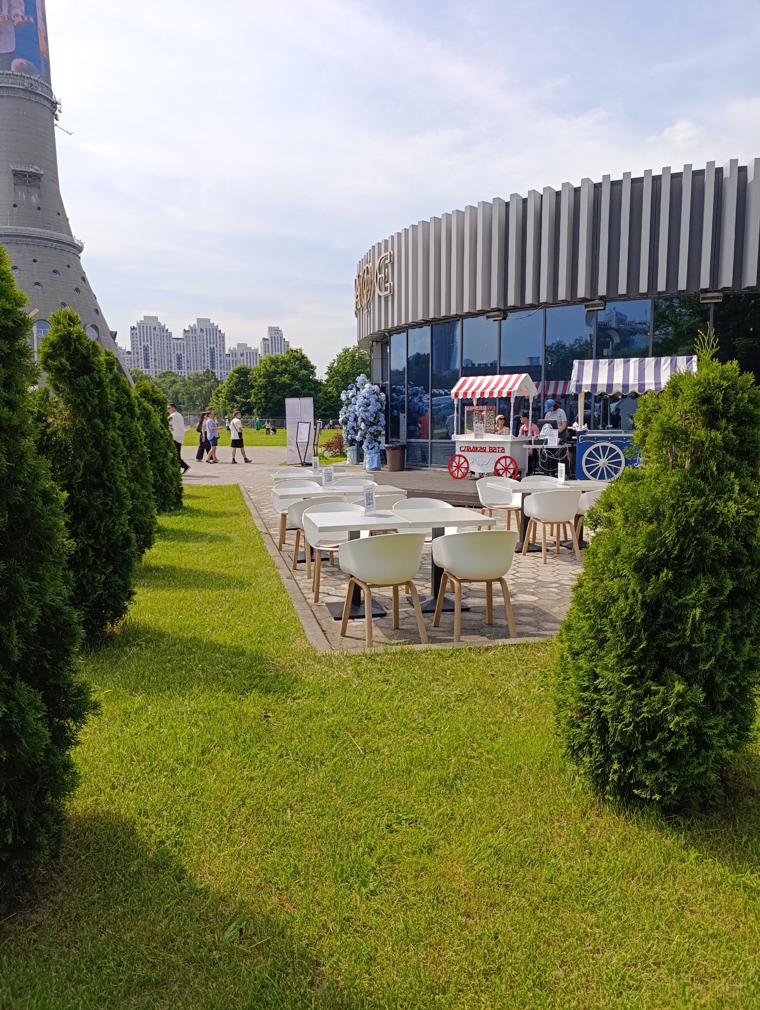 This image depicts a sunny urban park scene in Moscow, Russia, identified by the prominent Ostankino Tower visible on the far left. In the foreground, neatly mown green grass covers the ground, bordered by rows of trimmed evergreen shrubs on both sides. A paved area in the mid-ground features an outdoor cafe setting with multiple white tables and chairs.
Behind the cafe seating, a modern, circular-shaped building with a distinct facade of vertical grey and white panels on its upper section and large reflective glass windows below dominates the mid-ground. A light-colored logo, possibly "GE," is visible on the upper left side of the building's facade. In front of the building, two colorful vendor carts are present: one with red and white stripes is labeled "СЛАДКАЯ ВАТА" (Sweet Cotton Candy), and another with blue and white stripes. Several individuals are positioned near these carts, with one person in a striped shirt appearing to be an attendant at the blue cart. Other people are seen walking along a path further back to the left of the building. Large blue floral decorations are situated near the building's entrance.
In the distant background, beyond the Ostankino Tower, several large, modern residential buildings with light-colored facades and ornate rooflines are visible, silhouetted against a bright, partly cloudy sky. The scene is illuminated by natural daylight, casting clear shadows on the ground and under the tables.