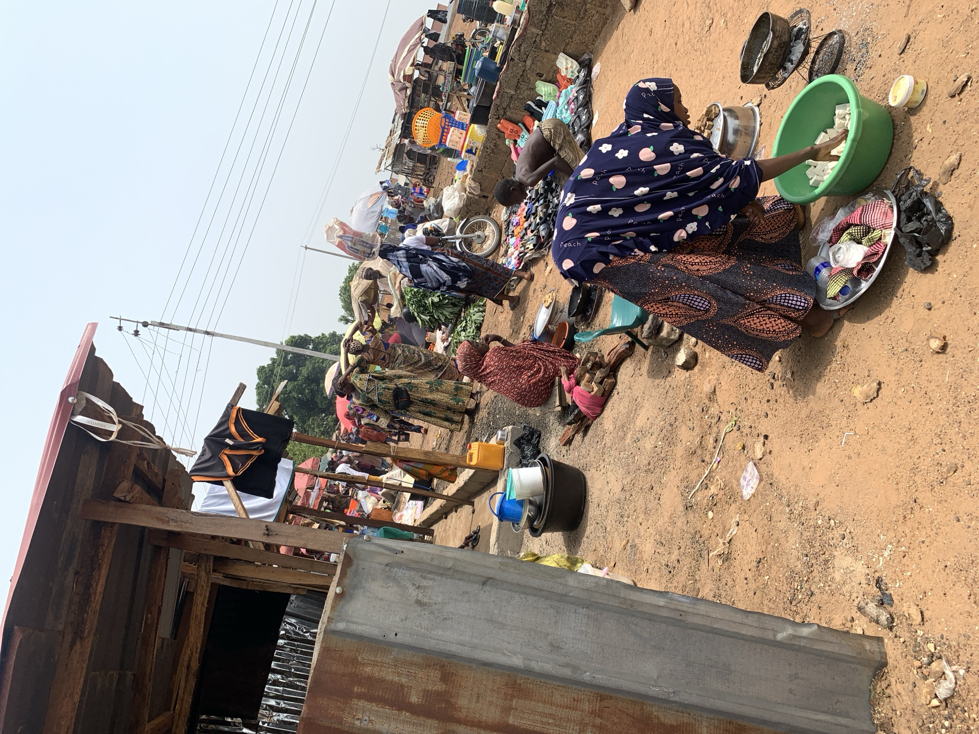 In Keffi, Nigeria, the scene depicts a bustling market on a dusty, sunlit day. Several women are seated on the ground, surrounded by goods and containers. One woman, wearing a navy blue headscarf and a patterned dress, is washing white cubes in a green plastic basin. Nearby, there are metal pots and dishes scattered on the ground. Other women are arranging or displaying their wares, which include leafy greens and various items spread out on mats. In the background, a cluttered market scene unfolds with makeshift stalls and a variety of goods for sale. Power lines stretch across the top left corner of the frame. A partially constructed or dilapidated building, made of wood and corrugated metal, is visible in the foreground, with clothing hanging from its structure.