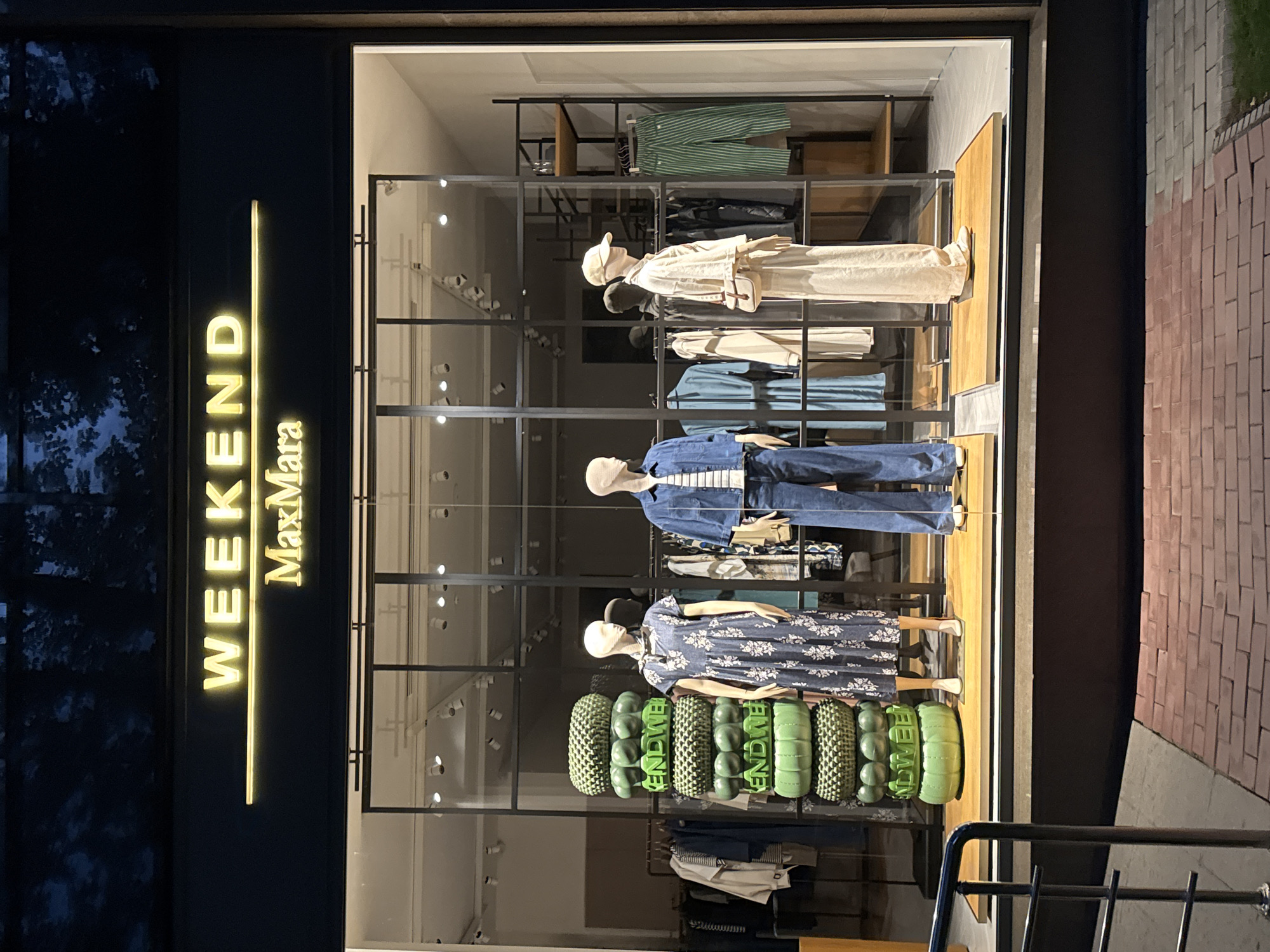 The image shows a well-lit storefront for "WEEKEND MaxMara" in Bishkek City, Kyrgyzstan, at night. The store features a large display window showcasing mannequins dressed in various outfits. In the foreground, a stack of green, textured decorative elements is visible. The mannequins are positioned in the center of the display, with one on the left wearing a floral print dress, another in the middle wearing a denim jacket and jeans, and a third on the right wearing a light-colored, loose-fitting outfit and a baseball cap. Behind the mannequins, racks of clothing are visible, including dresses, jackets, and striped pants. The storefront is framed by dark metal supports and illuminated by overhead lighting within the store. The exterior pavement is made of red brick.