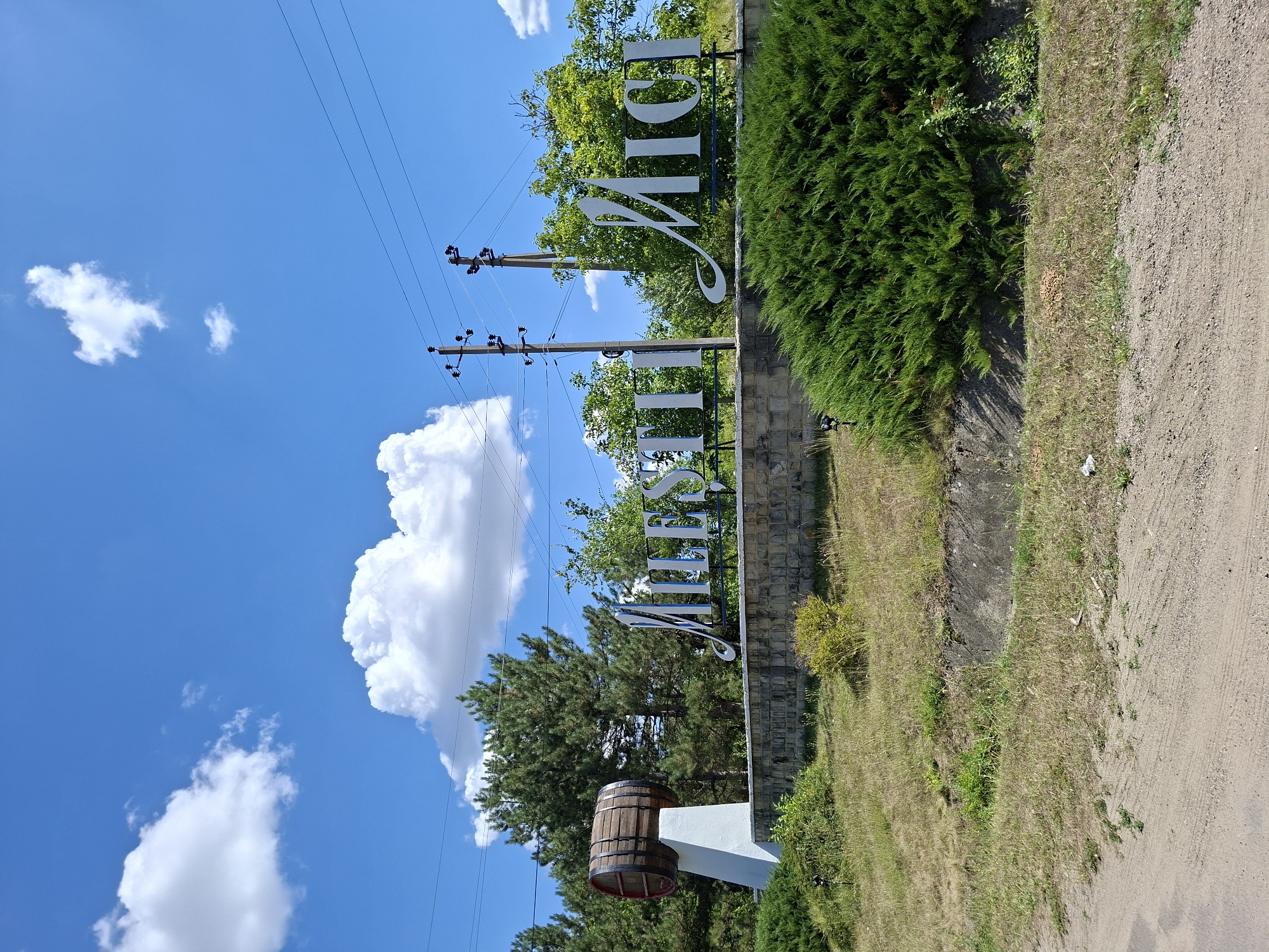 In Mileștii Mici, Moldova, a sign with large white, stylized lettering on a dark blue background announces the name of the town. The sign is situated behind a stone wall and in front of a dense evergreen bush. To the left of the sign, a large wooden barrel rests on a white pedestal. In the background, several tall pine trees are visible, along with other deciduous trees and shrubs. Overhead, a bright blue sky is dotted with fluffy white clouds. Electrical poles and wires are present above the trees. The foreground consists of a dirt road or path and dry grass. The overall scene depicts an entrance sign for the town.