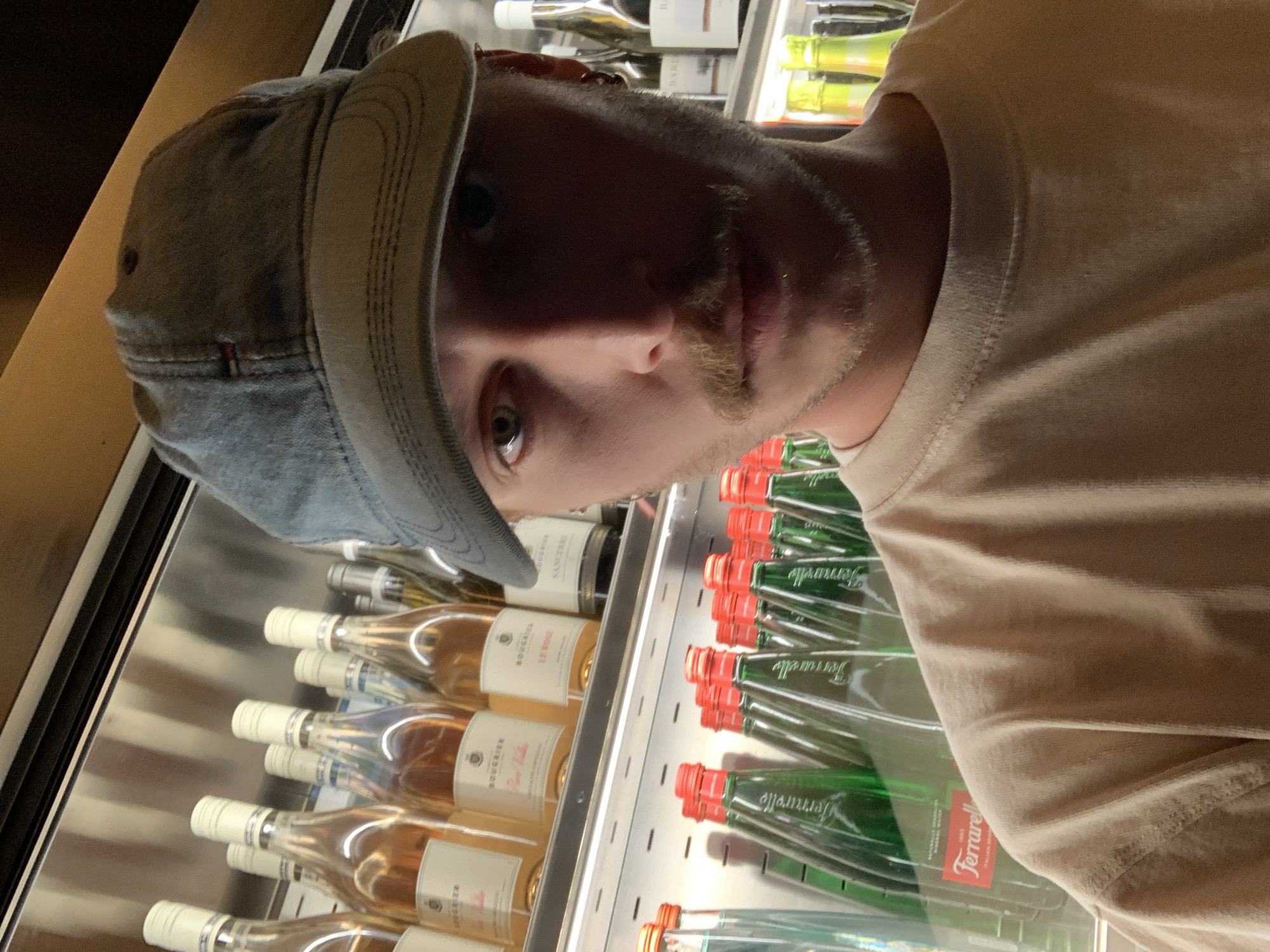 A man in a denim cap and a light tan t-shirt stands in front of a refrigerated display case. The case contains rows of wine bottles labeled "Rosé" and "Le Rose" and bottles of Ferrarrelle sparkling mineral water. The location is likely a grocery store in Tel-Aviv, Israel, as the water brand is commonly found there. The man appears to be looking at the contents of the display case.