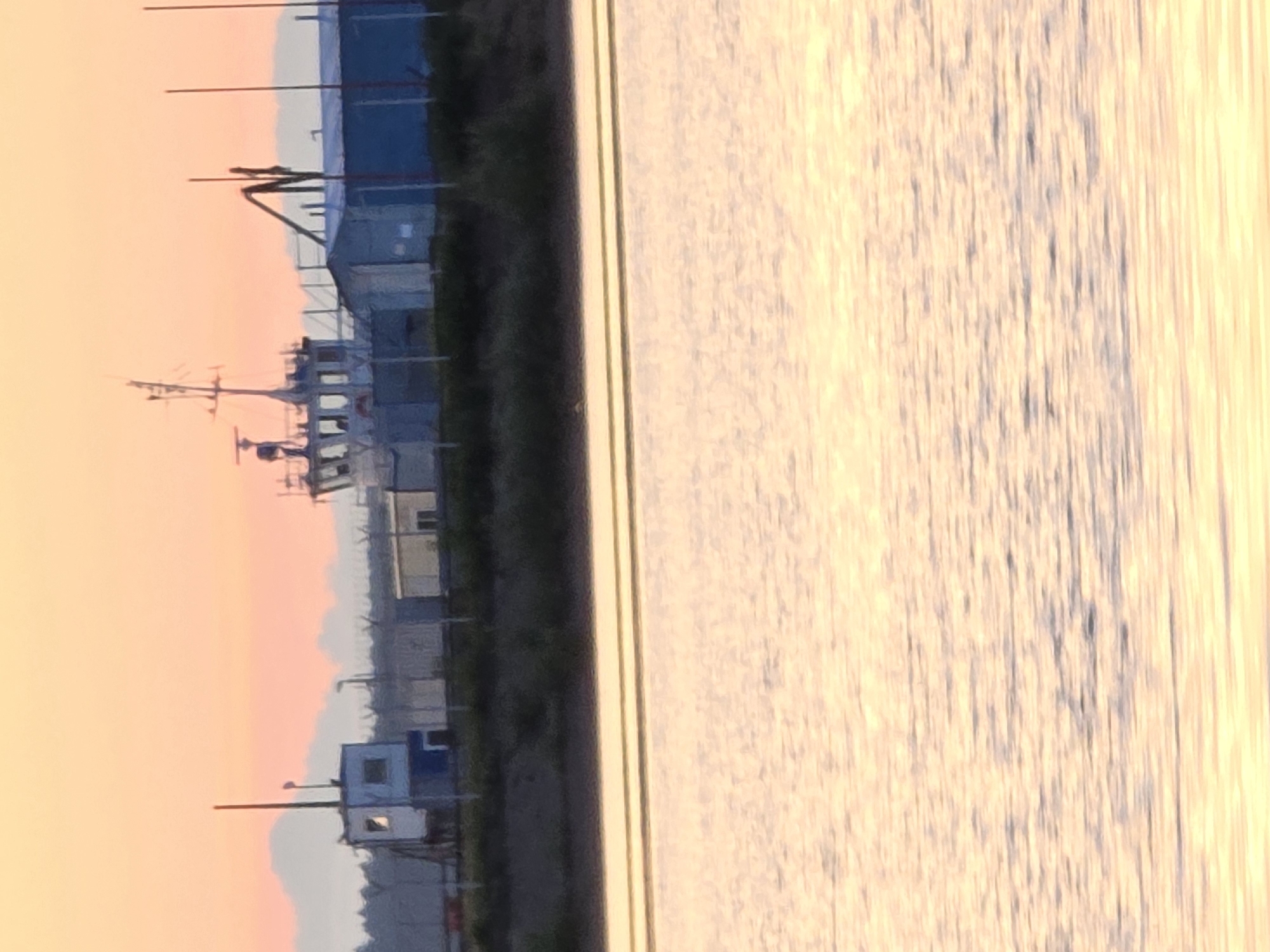 This image depicts a tranquil scene at either dawn or dusk along a body of water in Chaikovsky, Russia. The lower half of the frame is dominated by the calm surface of the water, showing subtle ripples and reflections of the sky's warm colors.
A narrow strip of dark land, possibly a shoreline with some vegetation, runs horizontally across the middle of the image, separating the water from the background. On this land, various structures are silhouetted against a softly lit sky. In the foreground on the left, there is a small blue and white cabin or security booth with a visible window, accompanied by a tall pole. Further along the shore, a line of what appears to be a fence or barrier can be seen, behind which are more indistinct buildings and industrial structures, some with blue elements. In the mid-ground, the distinct silhouette of a boat or ship with its masts and superstructure is visible, alongside what looks like a large industrial crane or piece of machinery.
The upper portion of the image is filled with the sky, displaying a beautiful gradient of soft orange, pink, and light blue, indicative of a clear sunrise or sunset. No specific activity or event is taking place, and no people are visible in the scene. The overall impression is one of calm and stillness, characteristic of a coastal or riverside industrial area at the quiet transition between day and night. There is no visible text in the image.