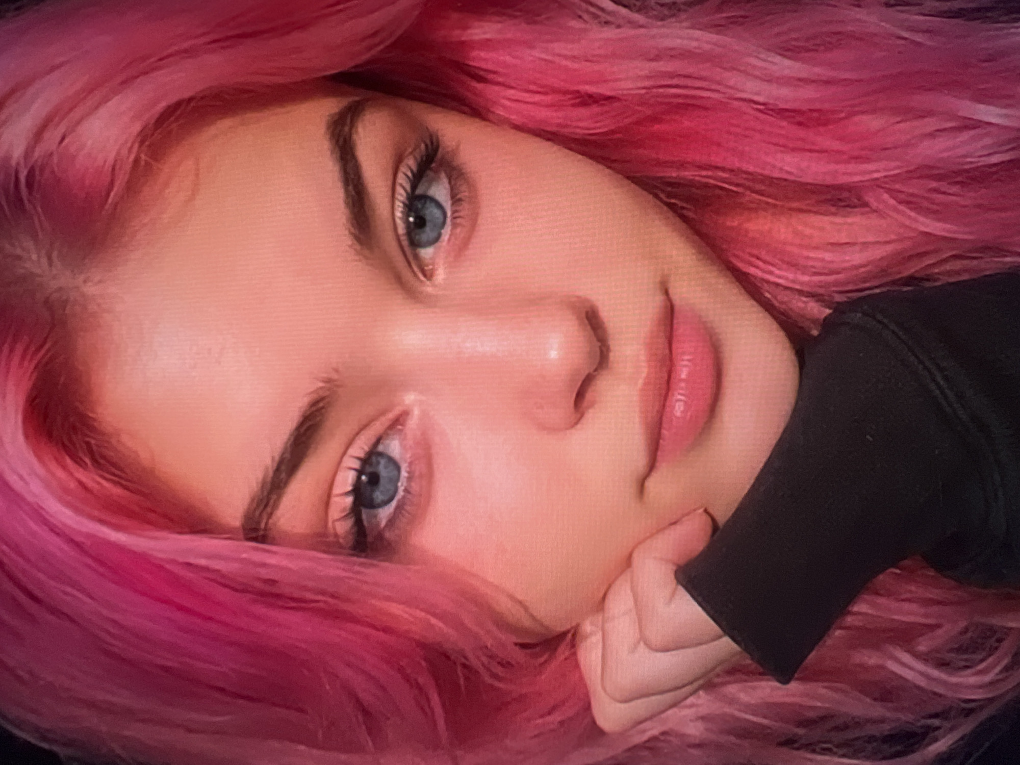 The image is a close-up, headshot of a young person with bright pink hair. Their face is turned slightly to the left, with their left hand resting on their chin, partially obscured by a black sleeve. The person has striking blue eyes, light skin, and full lips. The lighting is soft and warm, highlighting the texture of their hair and skin. The background is dark and out of focus, emphasizing the subject. The setting is not discernible from the image.