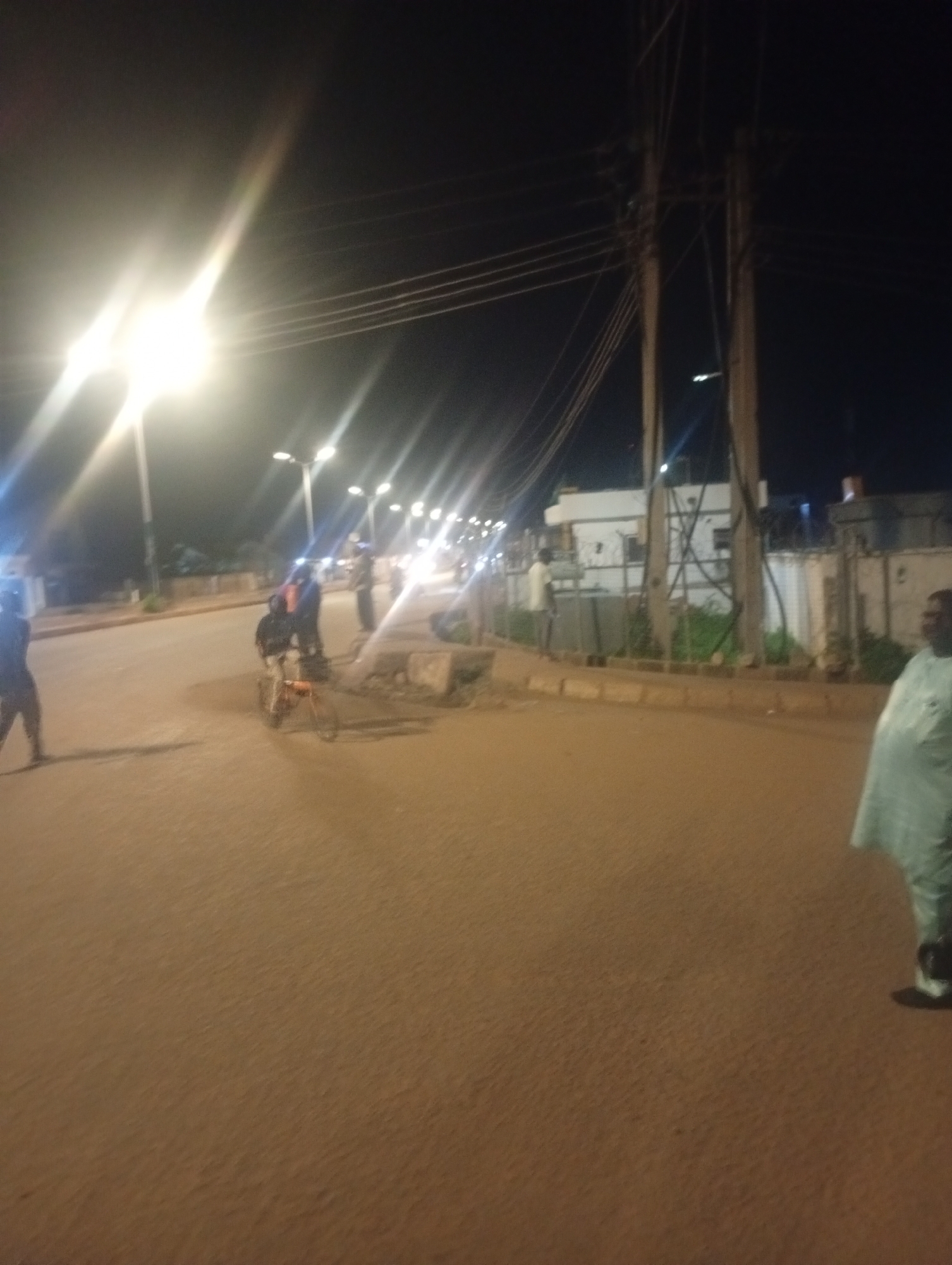 The image depicts a nighttime street scene in Bukuru, Nigeria, under the illumination of multiple streetlights and building lights. A person is riding a bicycle on the asphalt road, with a second individual standing behind them. To the right, another person is walking away from the camera, wearing a light-colored robe and dark shoes. In the background, there are buildings, a concrete curb, and overhead power lines. The overall impression is a busy urban environment at night.