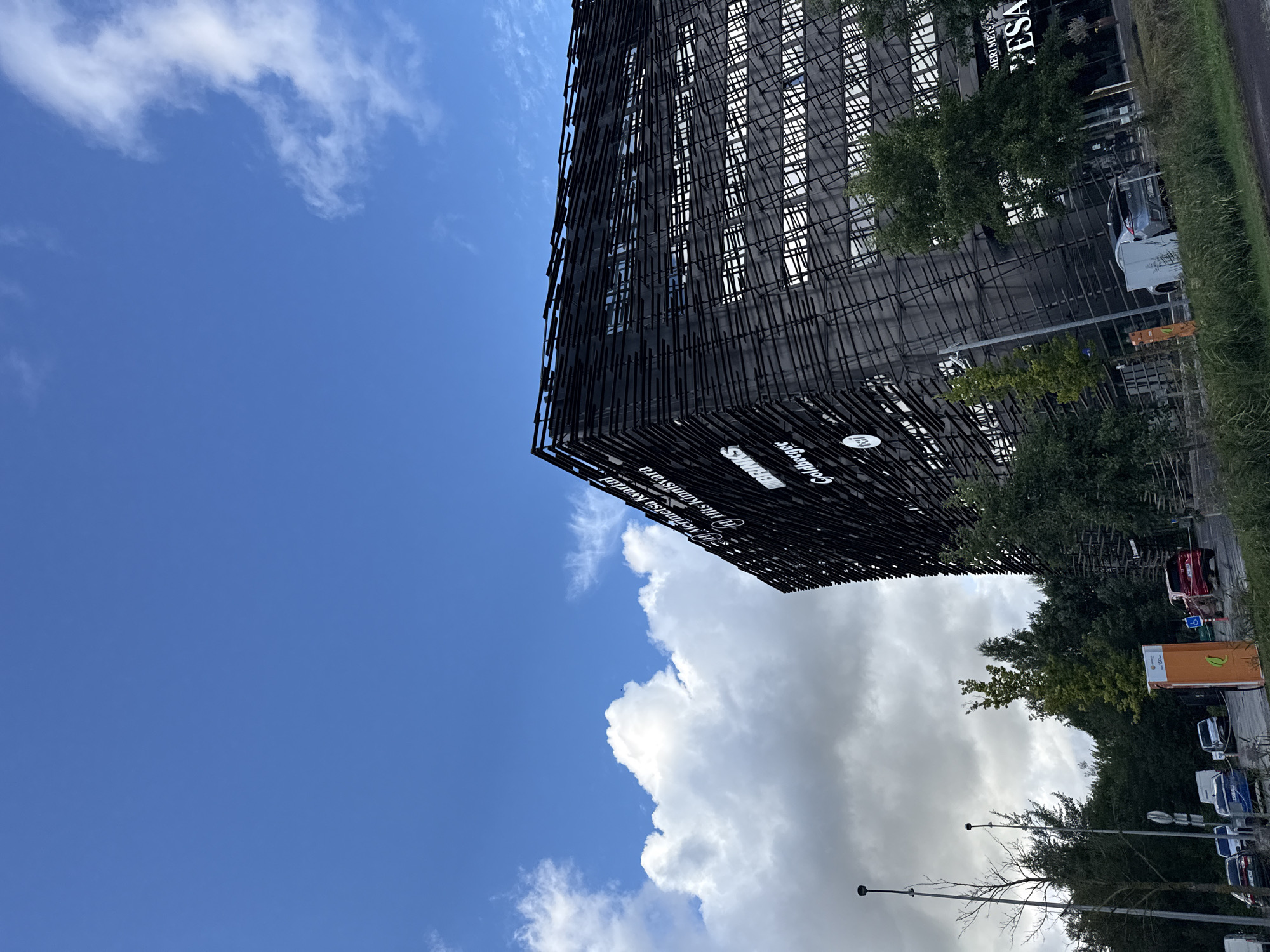 The image shows a modern black building with a distinctive facade, likely an office or commercial complex, situated in Tallinn, Estonia. The building's exterior is characterized by a pattern of horizontal and vertical dark elements, creating a textured and layered appearance. White lettering on the building indicates the presence of multiple businesses, including "MeriMetsa Kvartal," "Tils Kinnisvara," "BRINKS," and "Goldberger," suggesting it's a mixed-use development.
The sky above is a vibrant blue with scattered white clouds, indicating fair weather. In the foreground and to the left of the building, there are trees, some bare and others green, hinting at a season of transition. Several cars are parked in a lot or on a road in front of the building, including a red car and a silver electric vehicle. A bright orange charging station for electric vehicles is also visible. The overall scene depicts an urban or suburban environment with contemporary architecture and infrastructure.