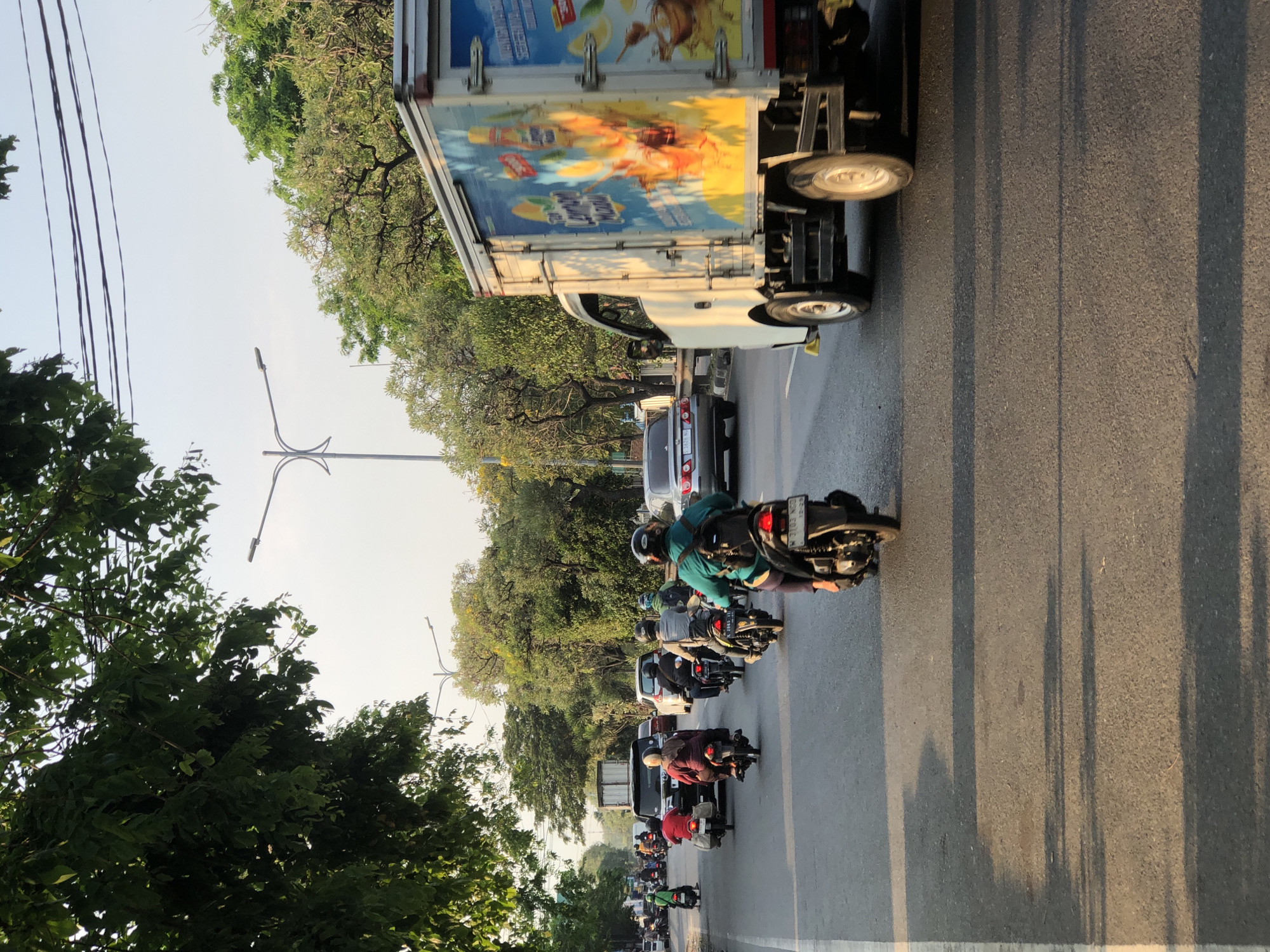 This media file shows a street scene in Kludan, Indonesia, with a procession of motorcycles and a truck. The subjects are primarily people riding motorcycles, wearing helmets and casual clothing. Objects include various motorcycles, a white box truck with colorful advertisements on its side, a few cars, and trees lining the street. The scene is a roadway with asphalt pavement and a white lane marker.
The activity taking place is a traffic jam or a slow-moving convoy of vehicles. The time of day appears to be late afternoon, suggested by the long shadows cast by the vehicles and the warm, golden light. The weather seems to be clear and sunny. The location cues are consistent with a tropical or subtropical environment, given the lush green trees. No specific emotions are clearly discernible from the distant figures, but the overall atmosphere suggests a busy, everyday commute.
Visible text is present on the side of the truck. It appears to be an advertisement for a beverage, with some text in what looks like Indonesian script. Some motorcycles also have license plates with text, though it is not clearly legible. The image is oriented vertically, with the sky at the top and the road extending downwards.