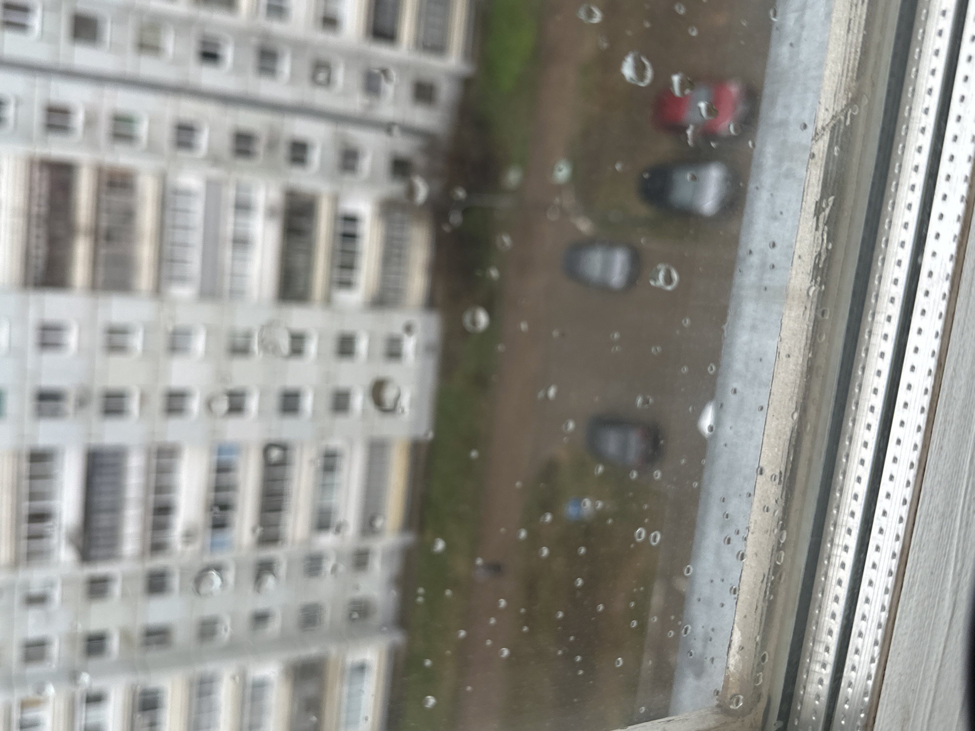 The image captures an overhead view from a building in Chaikovsky, Russia, looking down through a rain-splattered window. The immediate foreground is heavily covered with water droplets of varying sizes, which significantly blur the distant scene.
On the left side, a large, multi-story residential building, likely an apartment complex, is visible but out of focus. Its light-colored facade is characterized by numerous rectangular windows and balconies arranged in horizontal rows, typical of urban architecture.
To the right, beyond a blurry expanse of green ground and what appears to be an unpaved area or parking lot, several vehicles are present, also blurred. A distinct red car is seen towards the top right, and at least two dark-colored cars (possibly grey or black) are parked further down.
A segment of a window frame occupies the far right of the image. It is light-colored, possibly made of PVC or metal, and features a vertical track with a dotted texture, suggesting it may be part of a sliding window mechanism. The frame shows some signs of dirt or wear.
The presence of water droplets indicates that it is currently raining or has recently rained. The lighting suggests it is daytime, likely under an overcast sky, as there are no harsh shadows. The overall scene is static, depicting a quiet urban environment from an indoor perspective on a wet day. No people or visible text are discernible.