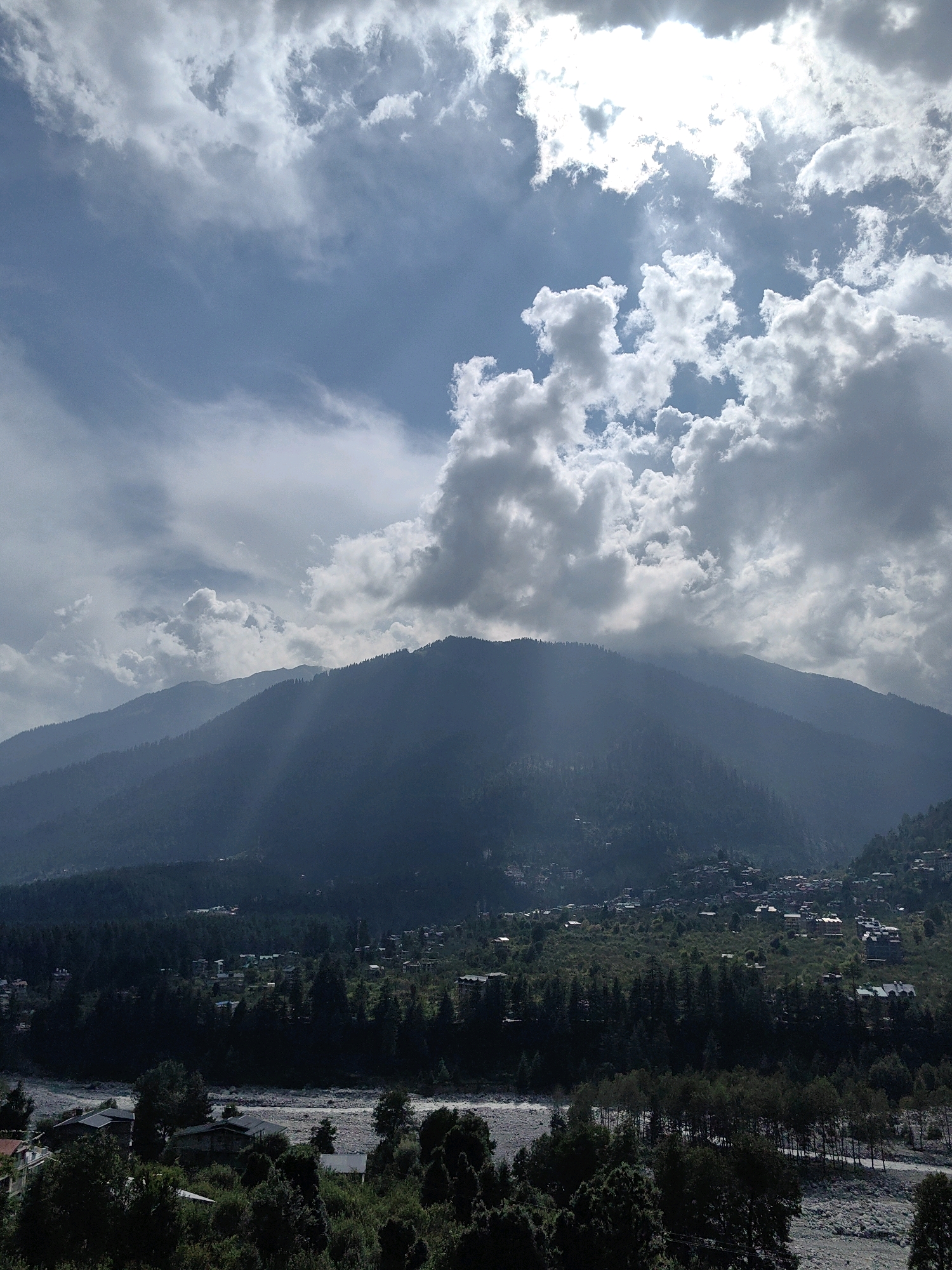The image depicts a serene mountain landscape in Vashisht, India. A village is nestled at the base of a large, tree-covered mountain. The sky is filled with dramatic clouds, with sunlight breaking through in several places, casting rays onto the mountain. A riverbed, partially covered with pebbles, runs through the foreground. The village itself appears to be composed of houses with red-tiled roofs, scattered amongst the trees. The overall impression is one of a peaceful, natural setting, with a small human settlement integrated into the vastness of the mountains.