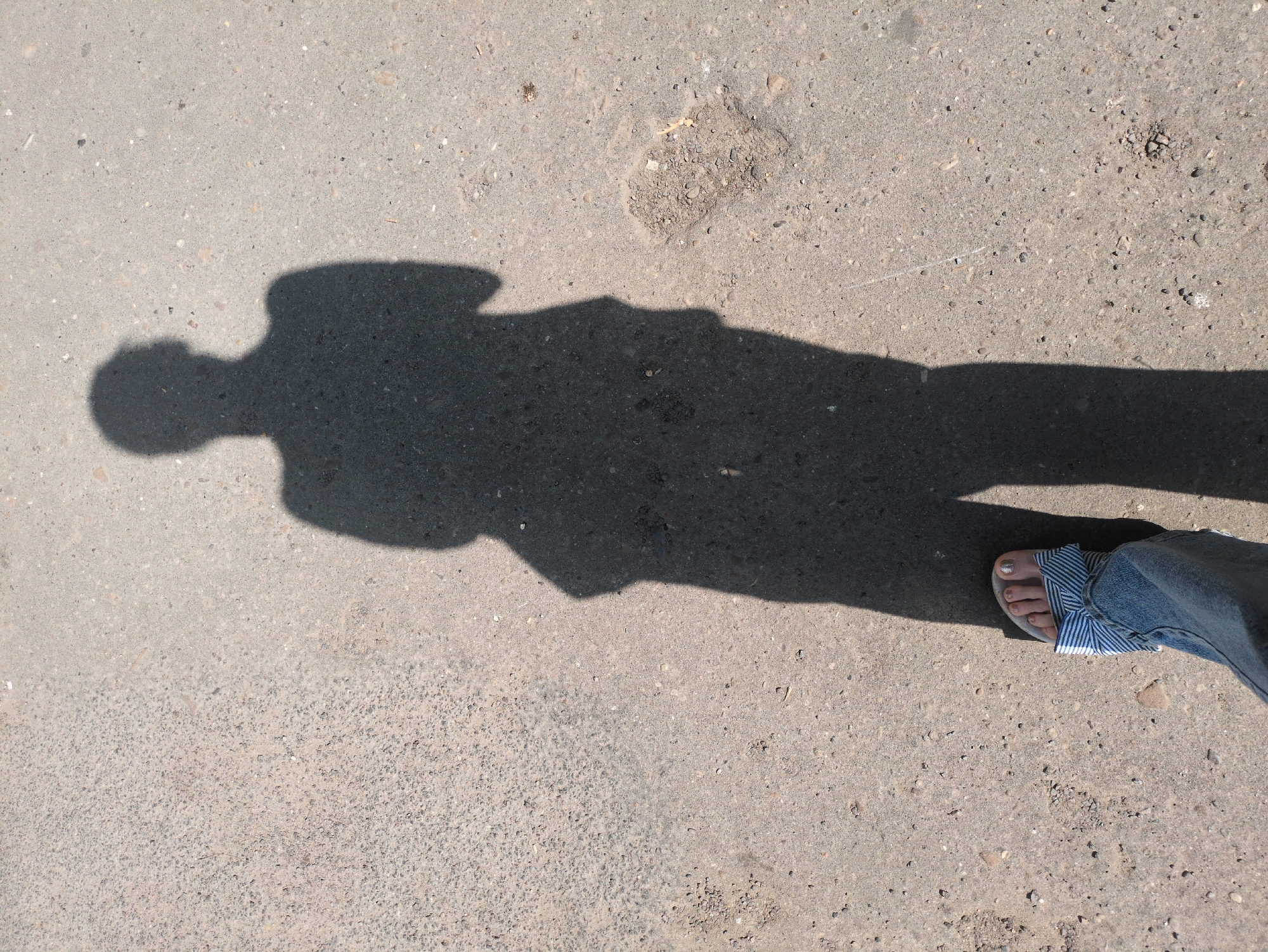 An overhead shot captures the shadow of a person standing on a gray asphalt surface. The shadow, elongated and dark, stretches from the lower left corner to the upper center of the frame. It clearly delineates the outline of a head, shoulders, torso, and legs. In the foreground, a person's left foot is visible, wearing a blue and white striped sandal and blue jeans. The asphalt surface is textured with small pebbles and is somewhat uneven, with some patches of dirt and debris scattered across it. The lighting suggests it is daytime. The location is Unknown, Unknown.