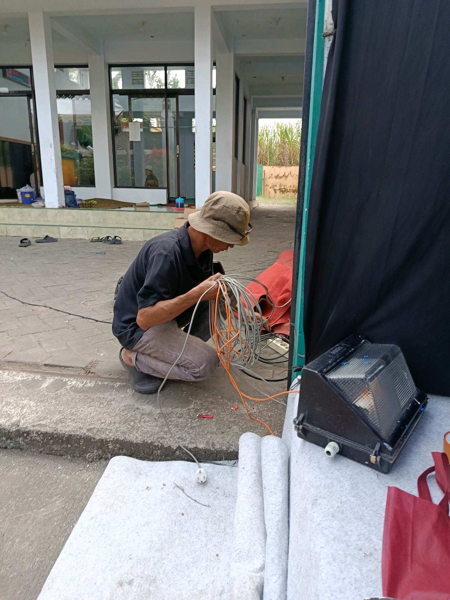 A man wearing a bucket hat and dark shirt kneels on a concrete path in front of a building with large windows. He is sorting through a bundle of orange and grey electrical cables. A black floodlight is positioned on a white fabric-covered surface to his right, with a red bag visible behind it. The building appears to be a public facility, possibly a mosque or community center, with several pairs of sandals on the ground outside. In the background, tall green plants are visible beyond a low wall. The scene is outdoors, with natural lighting. The location is the area in front of the administrative building of the Pondok Pesantren Modern Al-Fattah in Bululawang, Indonesia.