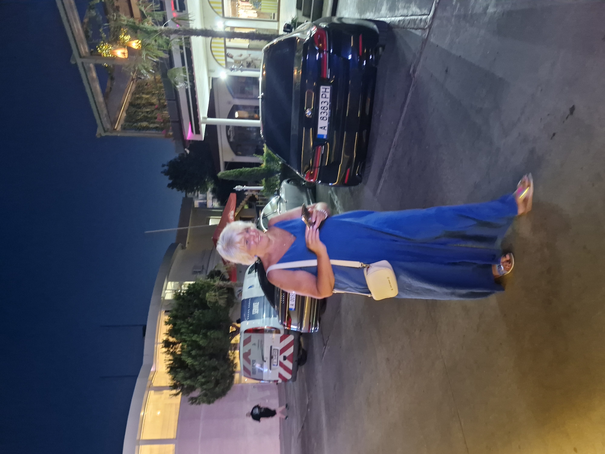 A woman in a blue dress and white sandals poses for a photo outdoors at night in Sveti Vlas, Bulgaria. She is smiling and holding a phone in her right hand. A small, beige crossbody bag is slung over her left shoulder. Behind her, a dark BMW car is parked, with a license plate reading "A 6383 PH." Further in the background, a white van with red and white striped markings and "SERVICE" written on it is visible. The scene appears to be in a commercial area, possibly near shops or restaurants, as suggested by the illuminated building facade in the background. The sky is dark, indicating it's nighttime, and the lighting suggests an urban or resort setting.