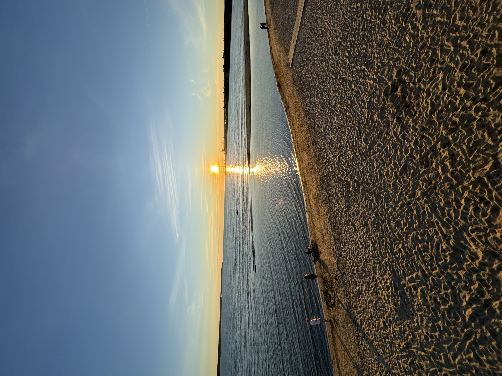 The image shows a beach scene at sunset, likely in Tallinn, Estonia, given the industrial skyline visible on the horizon across the water, which is characteristic of Tallinn's port area. The foreground is dominated by a sandy beach with the textured patterns of ripples and shadows cast by the low sun. Several small groups of people are present. On the left, two individuals stand near the water's edge, and a third is partially obscured. Further down the beach, two more figures are walking together. In the distance, on the right side of the beach, two people are walking away from the camera. The sea stretches out to the horizon, reflecting the warm hues of the sunset. The sun, a bright orb, is low in the sky, casting a shimmering path of light across the water. A thin strip of land with buildings and industrial structures, including cranes, forms the distant background. The sky is a gradient of blue, transitioning to warmer tones near the horizon.