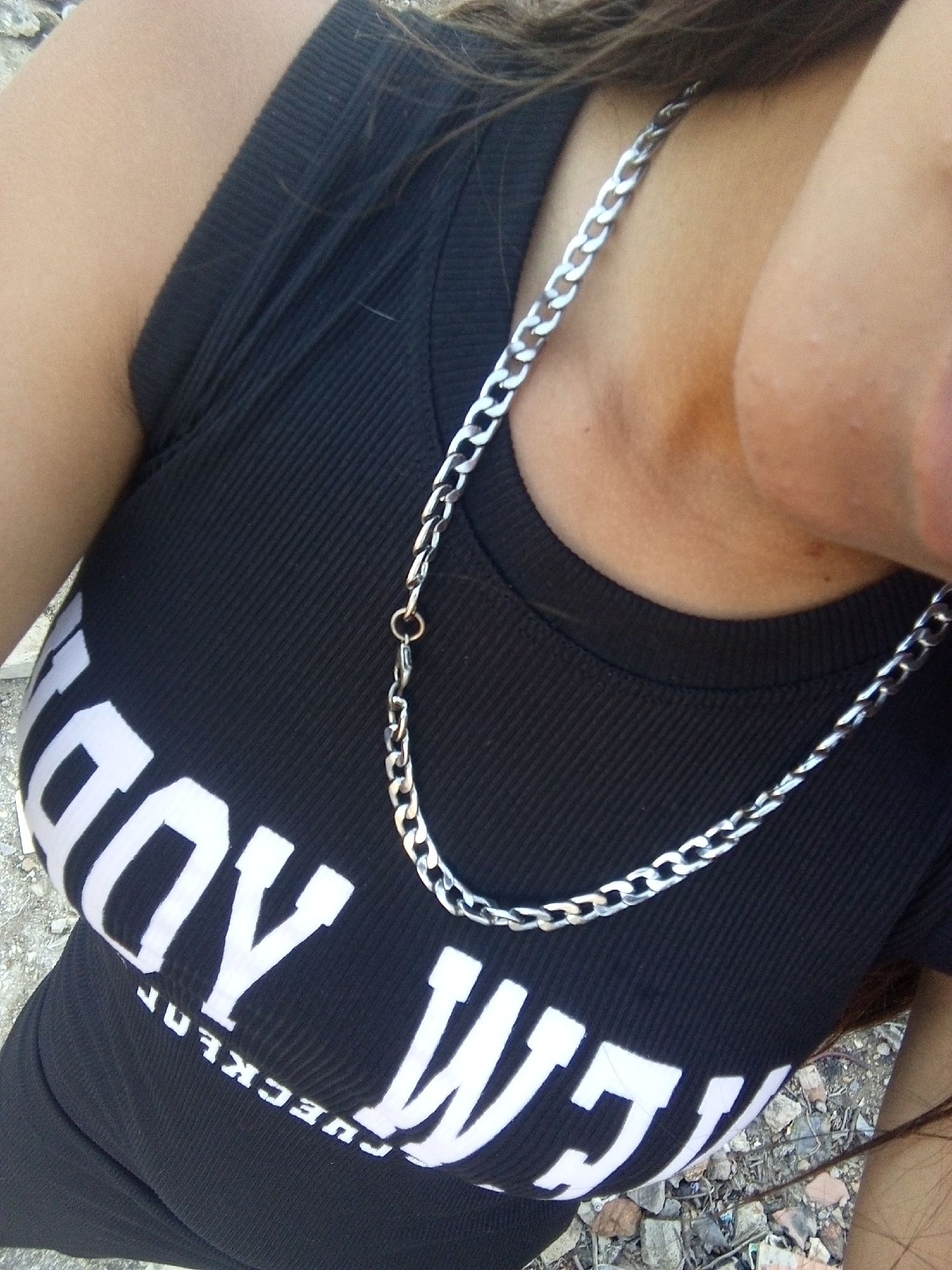 A photograph taken outdoors, presumably in daylight, depicts a person wearing a black, ribbed tank top with white lettering that reads "CHECKERBOY." A silver-colored chain necklace hangs around the person's neck. The environment appears to be a paved or rocky surface with scattered debris. The image's location in Juan Griego, Venezuela, is assumed but not explicitly evident in the picture.
