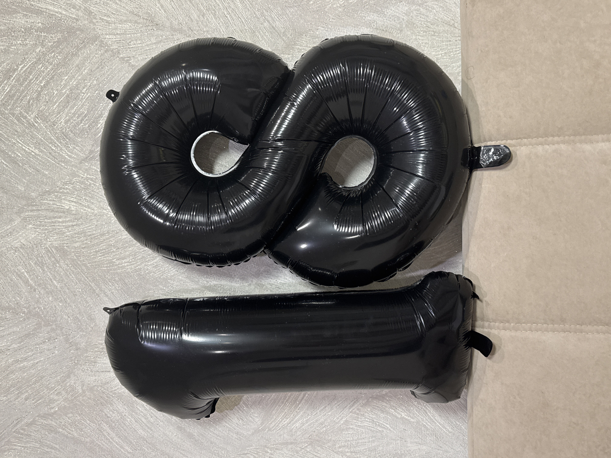 Three inflated black foil balloons are displayed in an indoor setting in Chaikovsky, Russia. Two of the balloons are shaped like the number '8' and are positioned horizontally side-by-side at the top of the frame. A third balloon, shaped like the number '1', lies horizontally below the two '8's.
The balloons are shiny and reflective, exhibiting typical creases and folds characteristic of metallic foil material. They are placed against two distinct background elements: a light-colored wall with a textured, brushed pattern on the left, and a beige or light brown upholstered couch or furniture piece on the right. The couch features visible stitching lines. Small black tabs, likely for hanging, are noticeable on the balloons.
The arrangement of the number-shaped balloons suggests decoration for a celebration, possibly a birthday or anniversary, where numbers like '18', '81', or '88' might be represented, although their placement isn't forming a single clear number. The scene is brightly and evenly lit, indicating an indoor environment. There is no visible text in the media file.