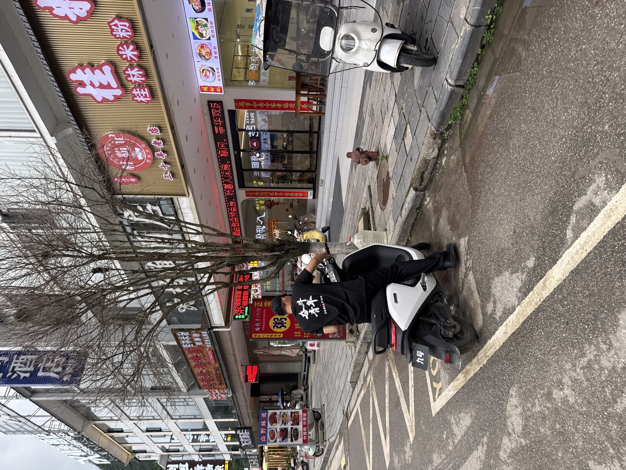 A man in black clothing and a baseball cap is lying on a white and black scooter parked along a street in Guilin, China. The scooter is parked next to a curb marked with white and yellow lines. Behind the scooter are several businesses with Chinese characters on their storefronts and signs, including restaurants specializing in Guilin rice noodles and other local dishes. A bare tree is visible near the scooter, and another scooter with a canopy is parked nearby. The street appears wet, and a fire hydrant is visible near the sidewalk.
