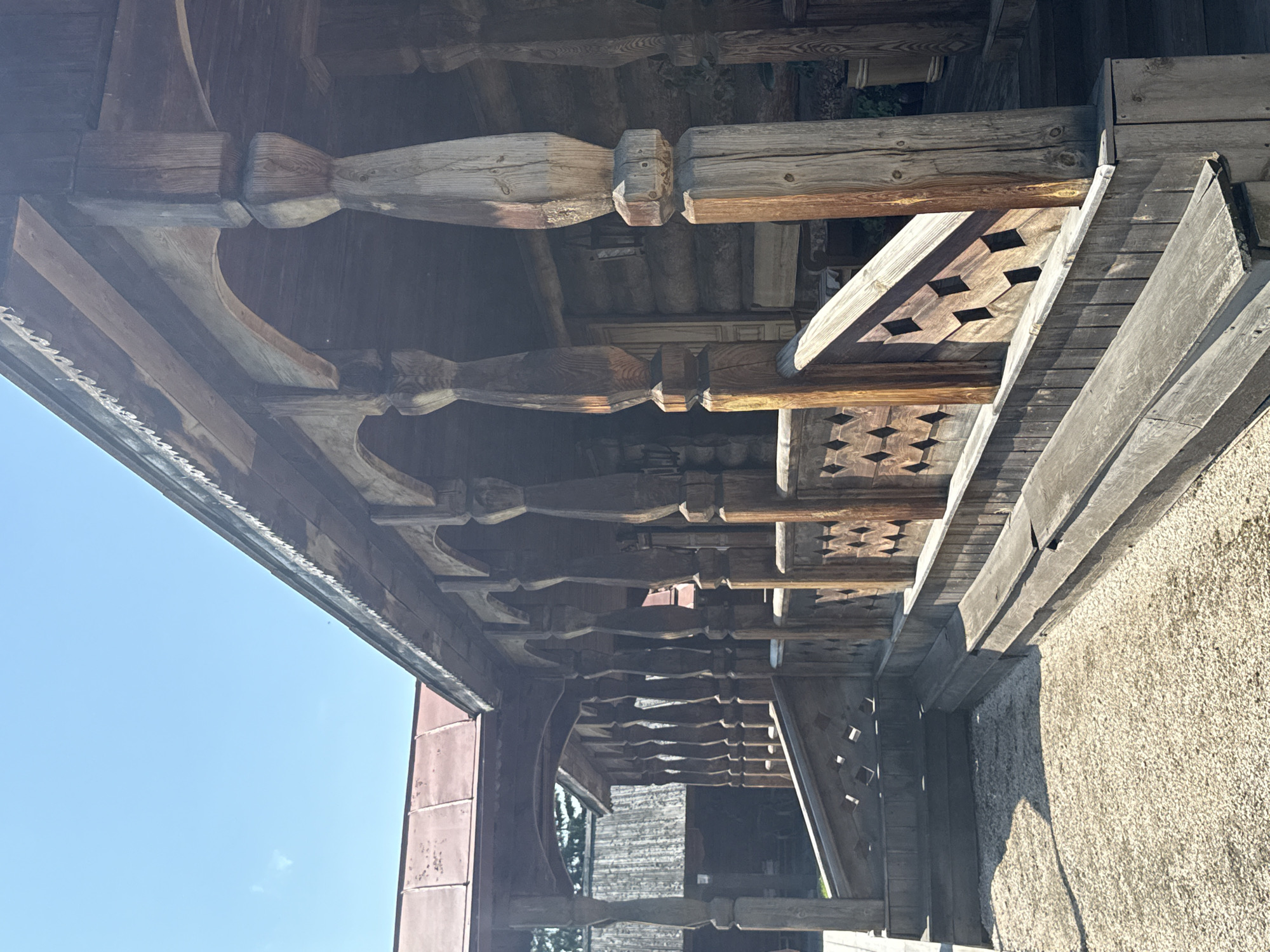 The image depicts the exterior of a traditional wooden log house with a covered porch, likely a historical exhibit or museum in Shushenskoye, Russia. The porch is supported by elaborately carved wooden posts, each with decorative turning and a hexagonal base. A wooden railing with a diamond-shaped cutout pattern runs along the porch. A wooden bench is situated in front of the porch railing. The ground in front of the porch is covered with light-colored gravel. The log walls of the house are visible behind the porch. The sky is clear and blue with a few wispy clouds. The overall scene suggests a warm, sunny day.