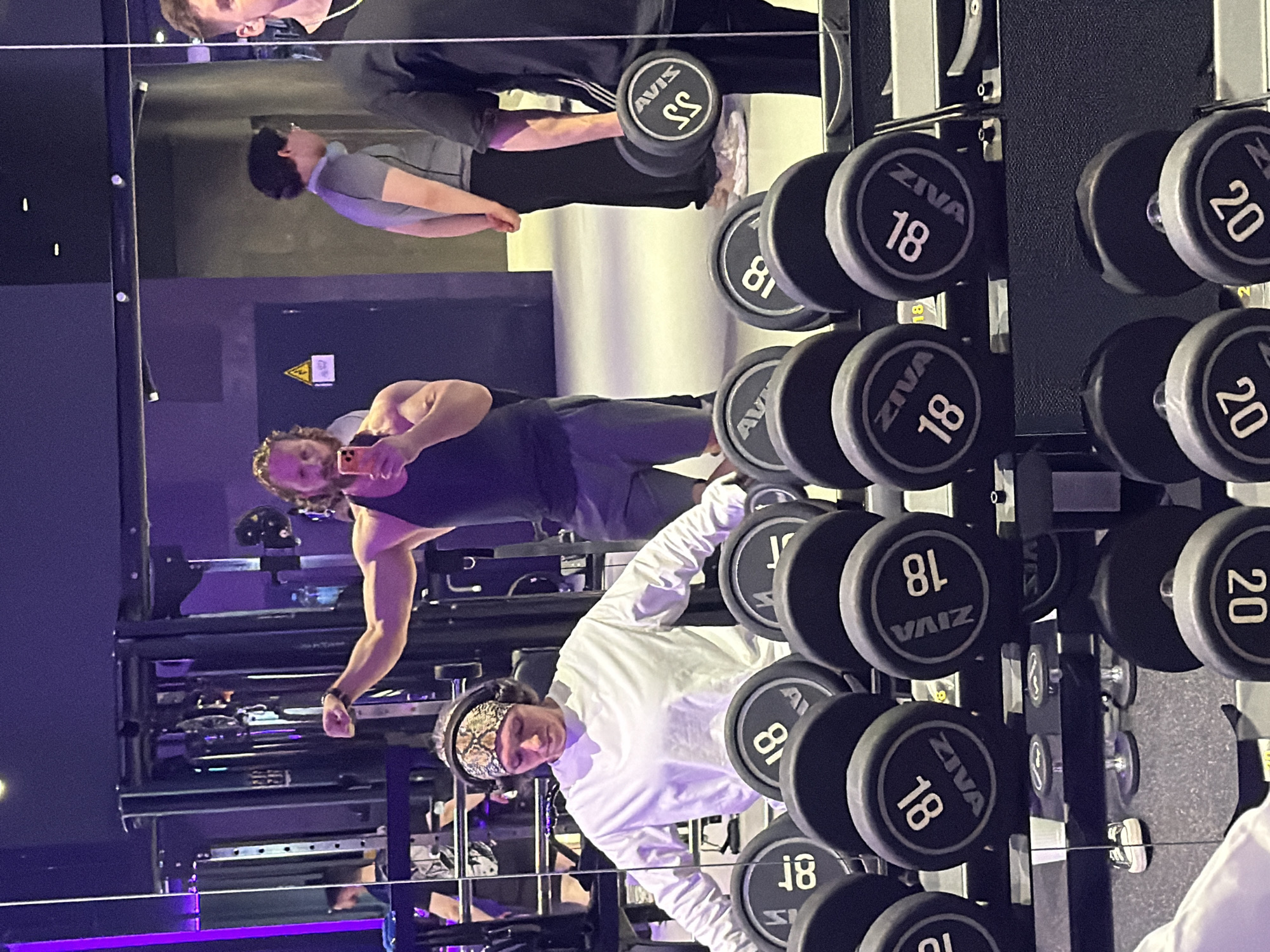 This image captures a vibrant, albeit somewhat dark, indoor gym scene, rich with reflections. The setting appears to be a modern fitness center, possibly in Saint Petersburg, illuminated by artificial lighting, which gives some areas a purple hue.
In the foreground, a person wearing a white long-sleeved shirt and a distinctive patterned bandana (possibly snake-skin or camouflage) stands near a dumbbell rack, looking downwards, perhaps preparing for an exercise or resting. Behind them, prominently reflected in a large mirror, is another man with curly hair, wearing a dark tank top. He is actively engaged in taking a selfie or video with a smartphone, holding it up while flexing his right bicep, showcasing his muscular physique. Further in the background reflection, another individual in a grey shirt is visible, and near the top, part of a fourth person.
The most prominent objects are the rows of black and grey ZIVA brand dumbbells on a rack. Visible weight numbers include "18" and "20". The reflection also shows "AVIS" on one of the dumbbells, likely due to a mirrored brand name or a different set. Other numbers like "55" and "81" are present, which are probably "20" and "18" seen upside down or in reflection. A yellow triangular warning sign with an exclamation mark is affixed to a wall in the background. The general activity is gym-goers working out or taking breaks, with one notable instance of self-documentation through a selfie.