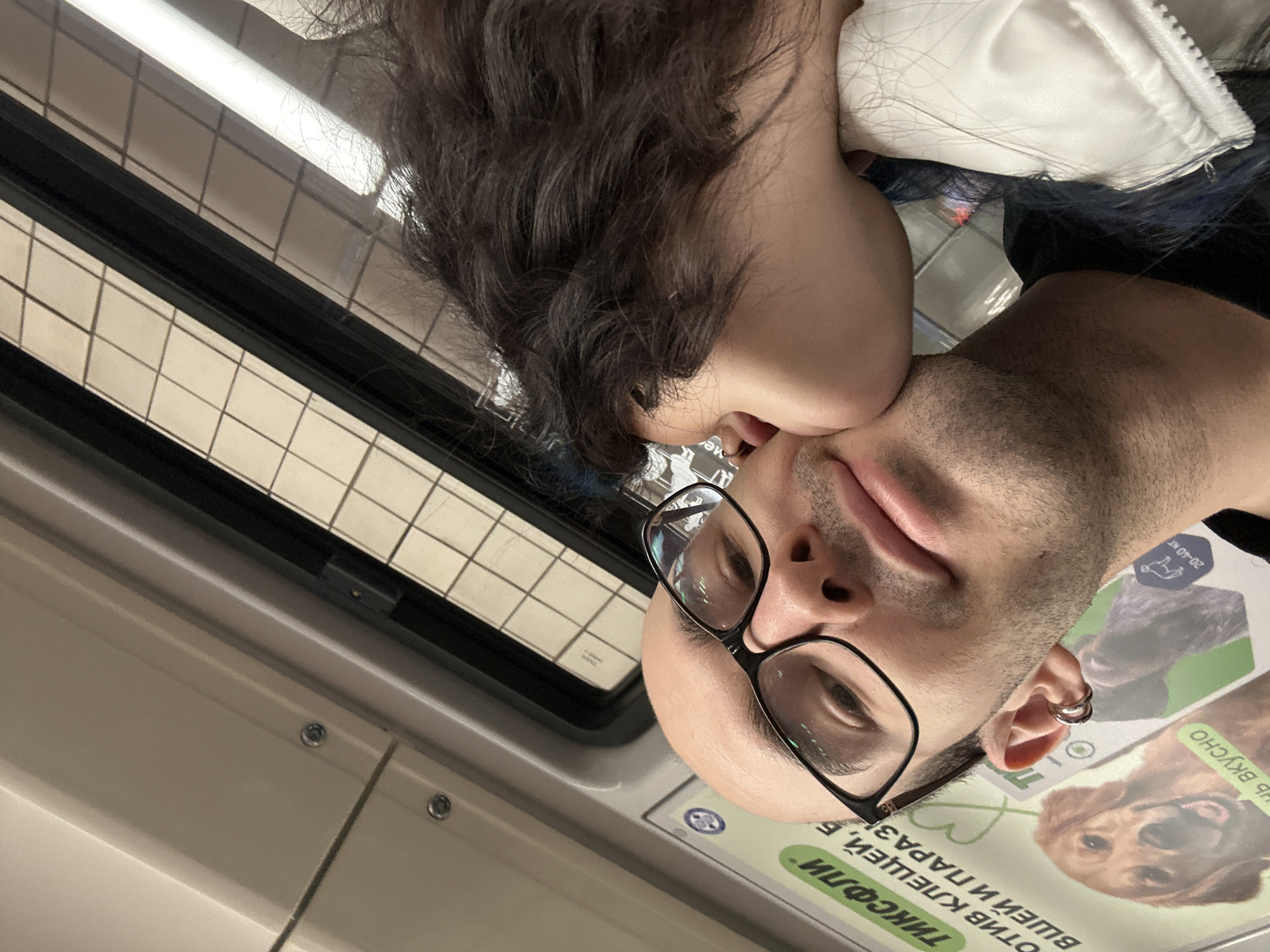 A close-up, slightly angled photo shows a man and a woman, possibly in a subway car in Moscow, Russia. The woman is positioned above the man, appearing to kiss him on the cheek. The man is wearing black-rimmed glasses and has short facial hair. He has multiple piercings in his ear. Behind them, there are advertisements in Russian, and the interior of what appears to be a subway car is visible. The window shows a tiled pattern from an unseen building outside the train. The angle is slightly canted, giving a casual, intimate feel.