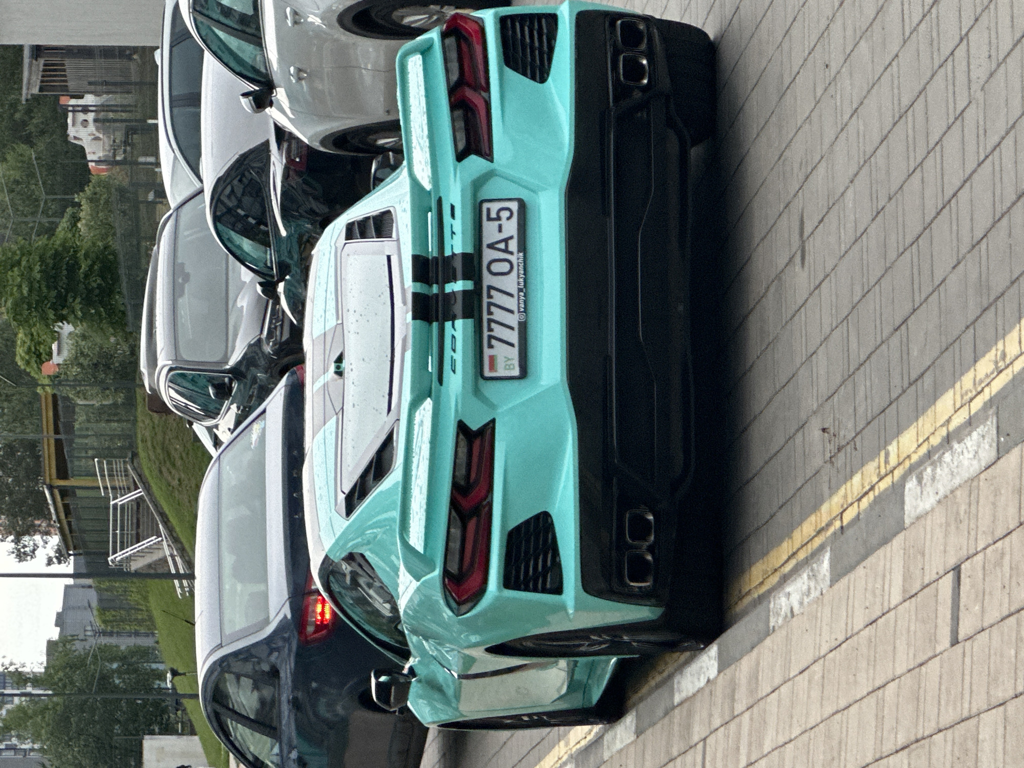 The image displays the rear view of a turquoise Chevrolet Corvette C8, parked on a paved surface next to a curb marked with yellow and white lines. The sports car features black twin racing stripes extending along its engine cover and rear spoiler. Its Belarusian license plate, "BY 7777 OA-5," is centrally affixed, with "@vanya_lukyanchik" printed below the registration number. The vehicle is equipped with quad black exhaust tips integrated into a black rear diffuser, and its red-tinted LED taillights are prominent, flanked by black mesh vents.
In the background, multiple vehicles are parked. Immediately behind and to the left of the Corvette is the rear of a dark-colored sedan with its left taillight illuminated. Further into the background, a cluster of parked cars, predominantly SUVs of varying shades, can be observed. The environment suggests an urban or suburban setting. To the left, trees and a distant building are visible. To the right, a grassy embankment features a set of concrete stairs with white railings leading up to a building with a yellow roof, situated behind a fence. The lighting indicates an overcast day. The license plate confirms the vehicle's registration in Belarus, specifically the Minsk Region (indicated by '5' in the regional code), which encompasses the Бараўлянскі сельскі Савет administrative unit. No individuals are present in the scene.