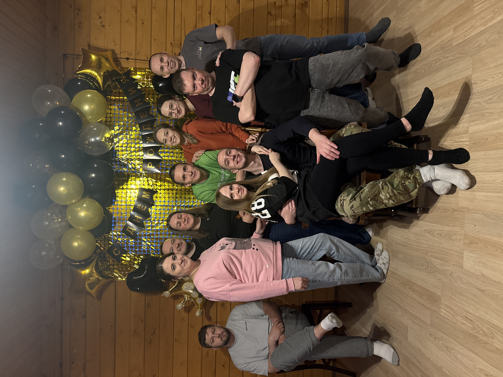 The image shows a group of twelve people posing for a photo inside a room. The backdrop includes gold and black balloons and a banner with the words "С Днём рождения!" which translates to "Happy Birthday!" in Russian. The room has wooden walls and a light wood floor. The people are arranged closely together, seated on chairs. The location is implied to be Chaikovsky, Russia due to the banner.