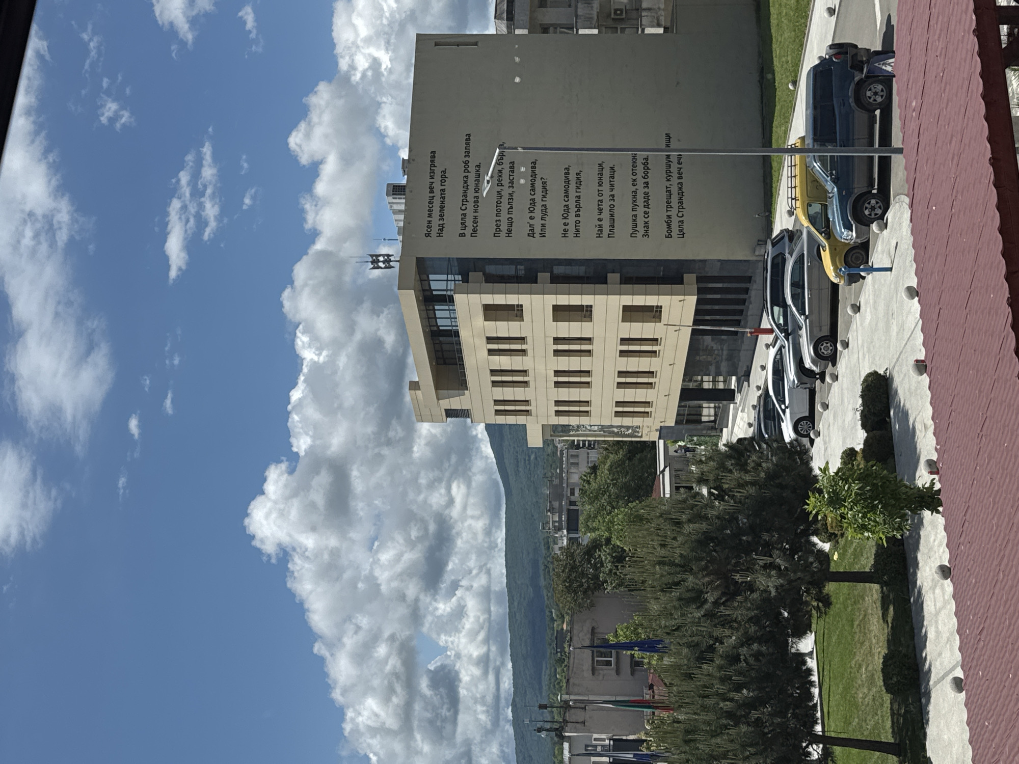 The image shows a view of a building in Tsarevo, Bulgaria, featuring a text from a Bulgarian poem on its side. The sky is partially cloudy, with the top portion a light blue color. A mountain range is seen in the distance behind the building. In front of the building, there is a row of parked cars and a grassy area with trees. A red roof is visible on the right side of the image, likely belonging to the building from which the photo was taken.
