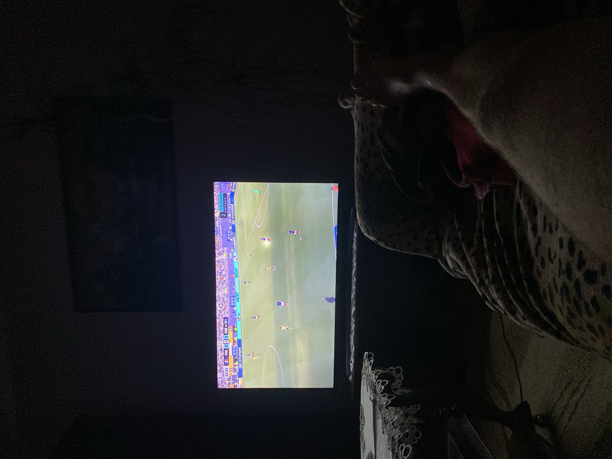 In a dimly lit room in Zastavna, Ukraine, a large flat-screen television displays a live soccer match. The score shows France 0 and Sweden 0 at 43:55. A person's legs and feet are visible in the foreground, draped in a leopard-print blanket, indicating they are likely sitting or lying down and watching the game. A small table with an ornate lace tablecloth is positioned to the left of the television. The room appears to be a living space, with the television being the central focus of attention.