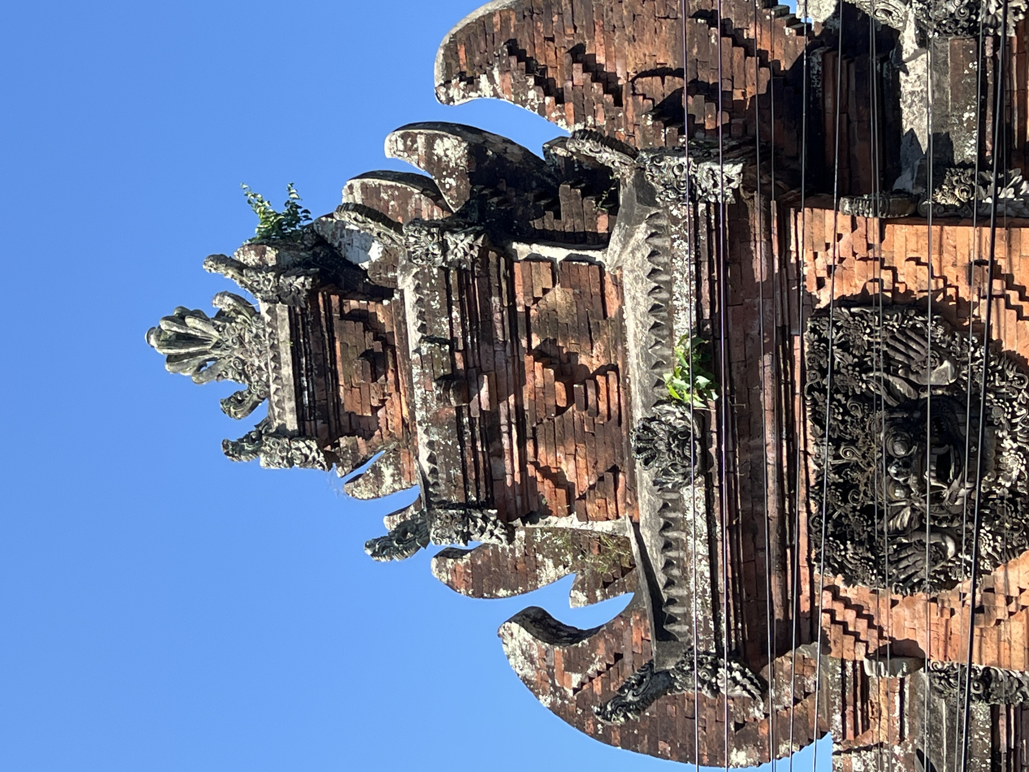 The image shows the upper portion of a traditional Balinese temple gate, specifically the Candi Bentar, located in Sukawati, Indonesia. The gate is constructed from reddish-brown brick, showing signs of age and weathering with patches of white moss and lichen. It features intricate stone carvings, including a fearsome demonic face on the lower section and decorative elements on the upper tiers. Several dark wires traverse the front of the gate, indicating the presence of electrical infrastructure. Small green plants are growing out of crevices in the stonework, adding a touch of nature to the weathered structure. The bright blue sky serves as a clear backdrop, with no other structures or people visible in the frame.
