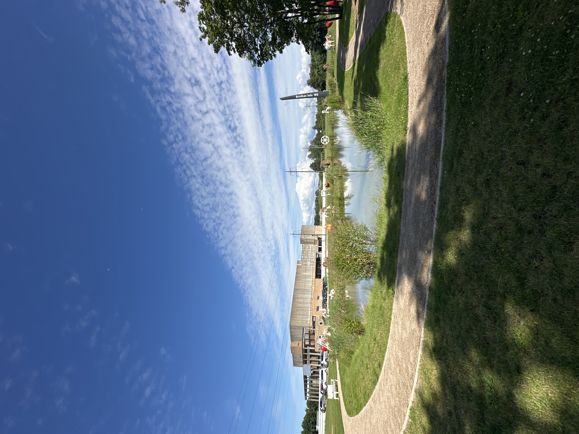 A wide-angle, eye-level shot captures a bright, sunny day in Liepaja, Latvia, with a vast blue sky dotted with wispy white clouds dominating the upper portion of the frame. In the foreground, a curved gravel pathway winds through lush green grass, partially shaded by the dappled sunlight. Beyond the path lies a serene pond, its surface reflecting the sky and surrounded by tall, green reeds.
On the far side of the pond, a large, industrial-looking building with a light-colored facade and dark columns stands against the horizon. In front of the building, a parking lot is visible with several cars parked. The scene is further enlivened by various outdoor art installations scattered around the pond and on the grounds. These include white sculptural figures, large red sphere-like objects, and a prominent black, tall, narrow sculpture with white text that reads "Brīvības iela 185." To the right of the pond, a mature tree with dark green foliage stands, and further back, a white cow sculpture is visible.
The overall atmosphere is peaceful and open, suggesting a public space or park, possibly an outdoor exhibition area. The presence of the building and the power lines in the background indicate an urban or semi-urban environment within Liepaja.