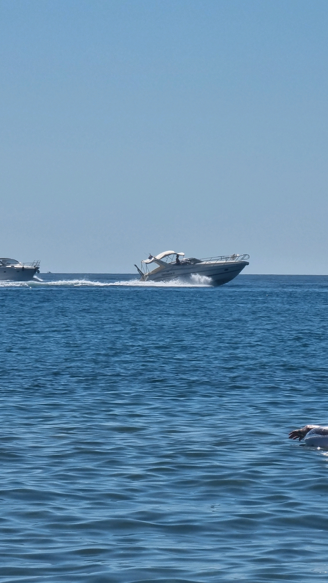 The image depicts a sunny day on the sea with a clear blue sky. In the mid-ground, two motorboats are active on the water. The boat on the right is a white speedboat with a canopy, moving at speed and creating a significant wake. A person is visible at the helm of this boat. To its left, another white motorboat is partially visible, also moving and leaving a wake. In the foreground, on the right side of the frame, a person's arm and hand are visible resting on a white inflatable float in the calm blue water. The scene is likely set in the coastal waters near Castelló d'Empúries, Spain, given the characteristic blue sea and sky.