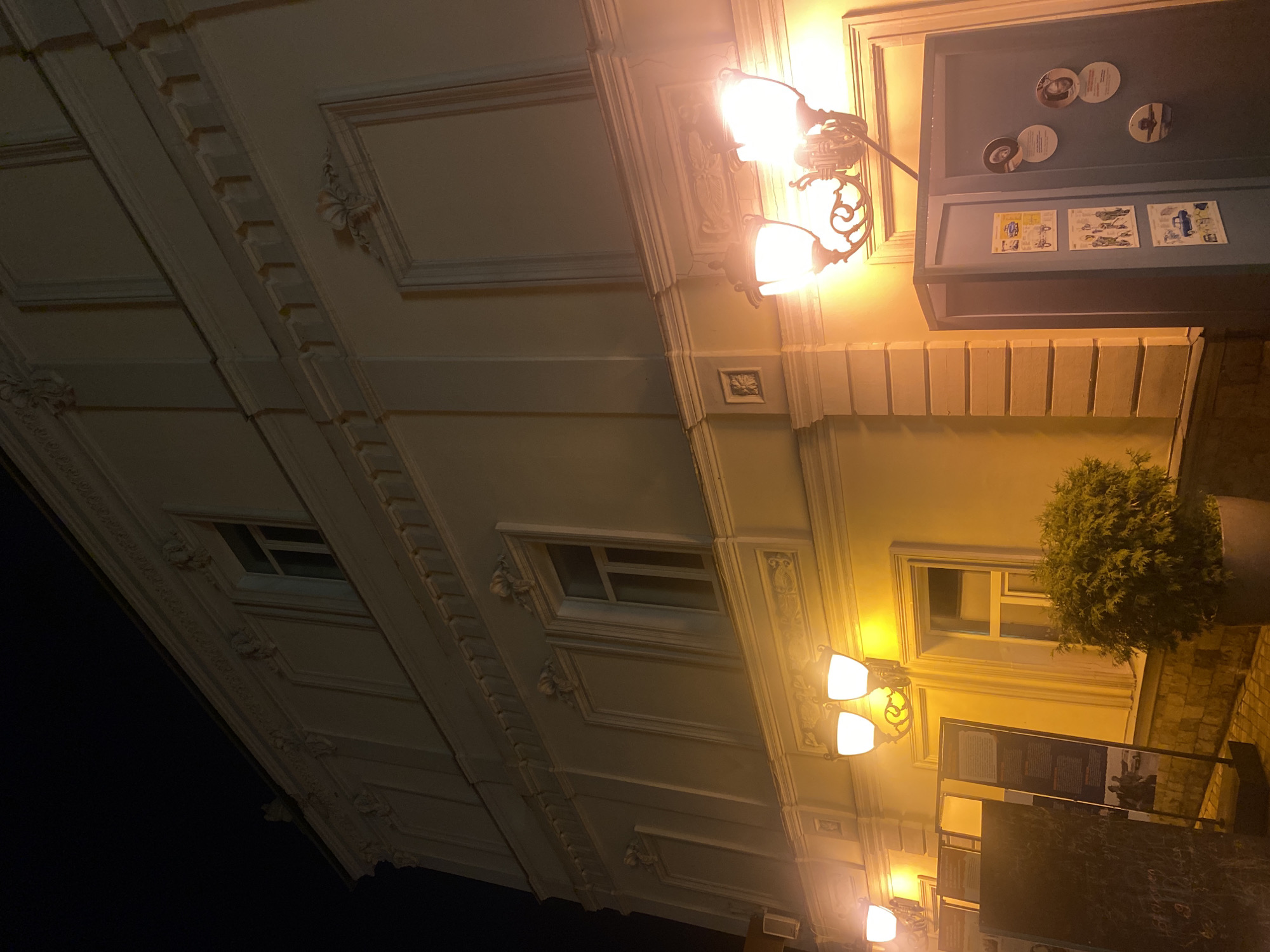 The image shows an ornate building facade at night. The building has detailed decorative moldings, cornices, and window frames. Two vintage-style wall sconces with warm, glowing bulbs illuminate the facade. One sconce is a double light fixture, and another is a single light fixture further down. The facade is painted a pale yellow.
In the upper right portion of the image, a dark blue recessed display case is visible. It contains several round coasters, each with a black and white image or text. Below these coasters are three rectangular cards, also featuring images and text.
Further to the right, a large potted evergreen plant with dense foliage is situated next to a window with a white frame. The window appears to be a modern style with horizontal panes.
Below the window and the plant, a brick-patterned wall is visible. A dark, frame-like structure with text and images is also present, partially obscuring the lower part of the building. The overall scene suggests an exterior view of a building, possibly a cafe, restaurant, or historical establishment, in an urban setting at night. The lighting creates a warm and inviting ambiance. The image is oriented such that the building facade appears to be stretching upwards and to the left. There is no clear indication of weather, but the darkness implies nighttime.
There is visible text on the items within the display case, but it is too blurry to read.
