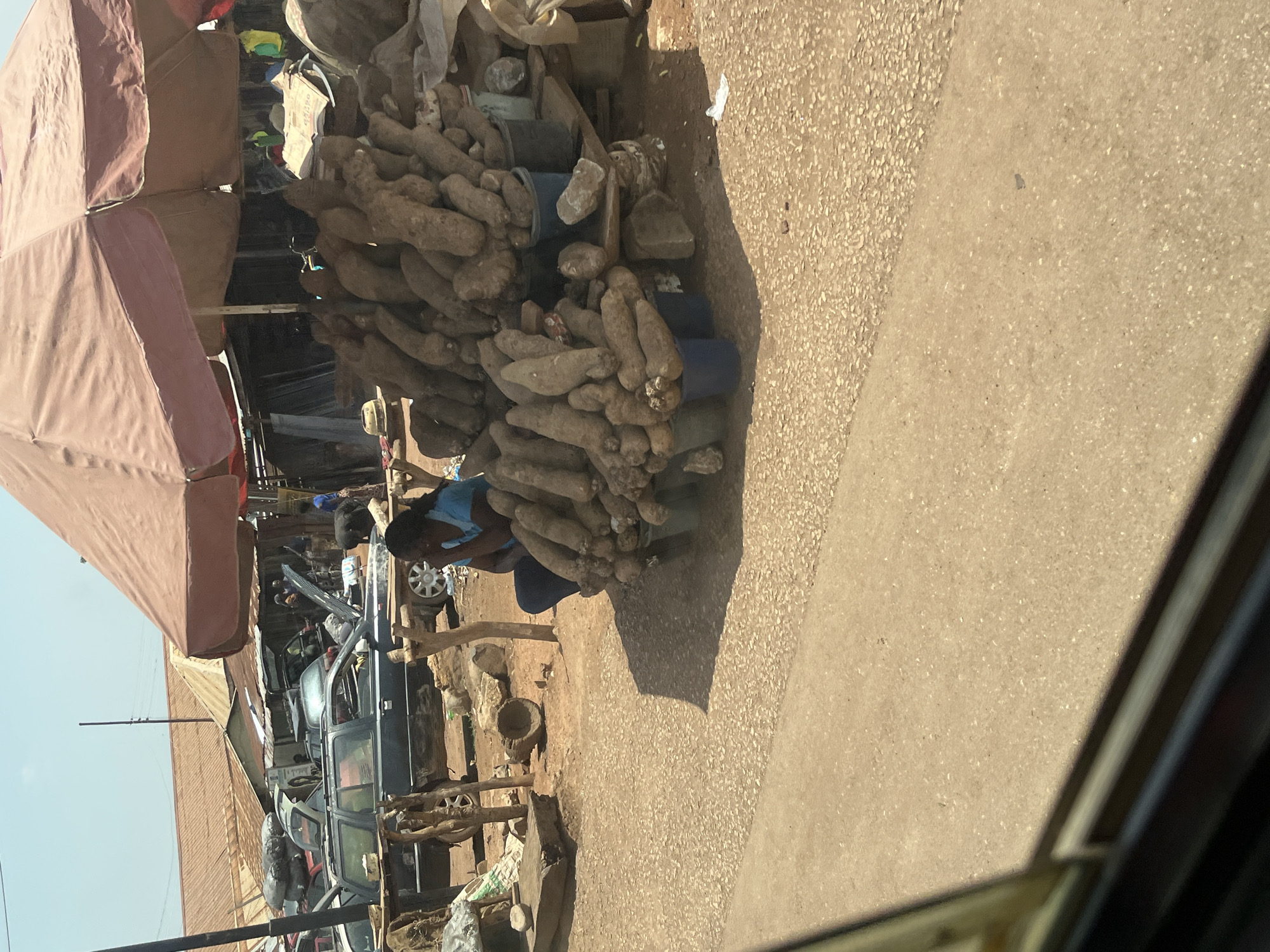 The image depicts an outdoor market scene in Karshi, Nigeria, under bright daylight. The main subject is a large, tall pile of yams or similar root vegetables, prominently displayed on the ground to the right of a paved area. A person wearing a blue top is seen sitting or crouching beside the yam pile, likely a vendor tending to their goods.
To the left, a large, reddish-brown market umbrella provides shade, extending over an unseen portion of the market. In the background, beyond the yam stall and umbrella, several vehicles are visible, some appearing to be parked or undergoing repair, with open hoods and visible car parts. Other people can be seen in the mid-ground, moving among the cars and what appear to be other market stalls or structures.
The ground is a mix of light-colored, speckled pavement and dusty earth. Strong shadows are cast by the market umbrella and other objects, indicating a sunny day, likely during the late morning or early afternoon. The setting suggests a bustling commercial area, characteristic of a local market in West Africa. No discernible text is visible within the image.