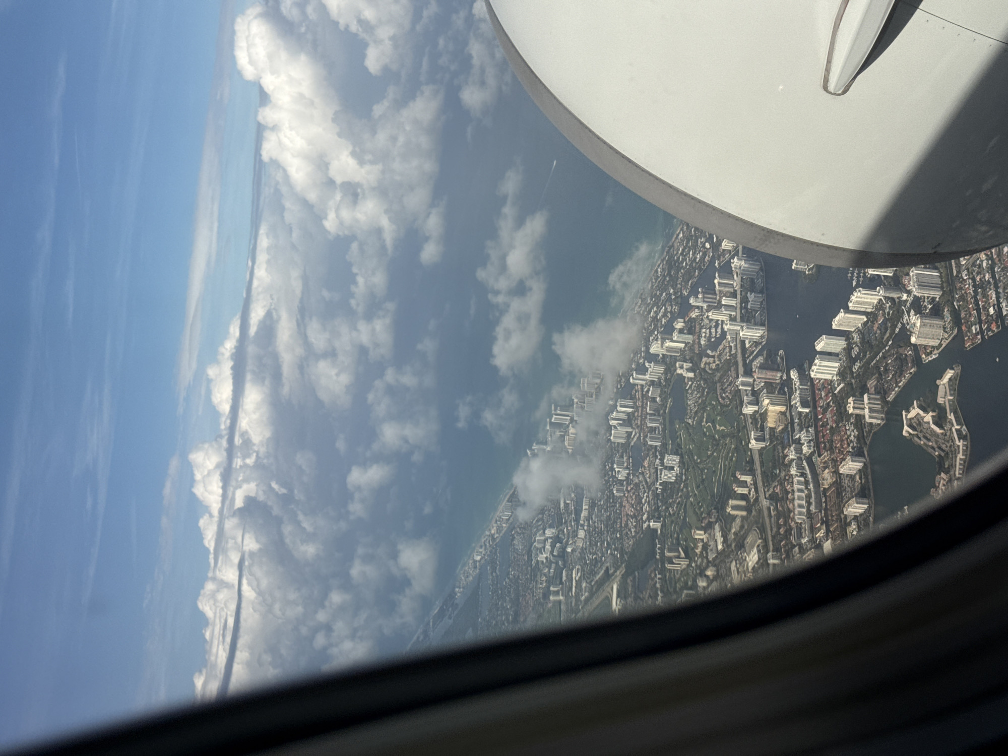 View from an airplane window over North Miami Beach, United States of America. Visible are a white airplane wing flap, sky with cumulus clouds, coastline, intracoastal waterway, residential and commercial buildings, and green spaces.